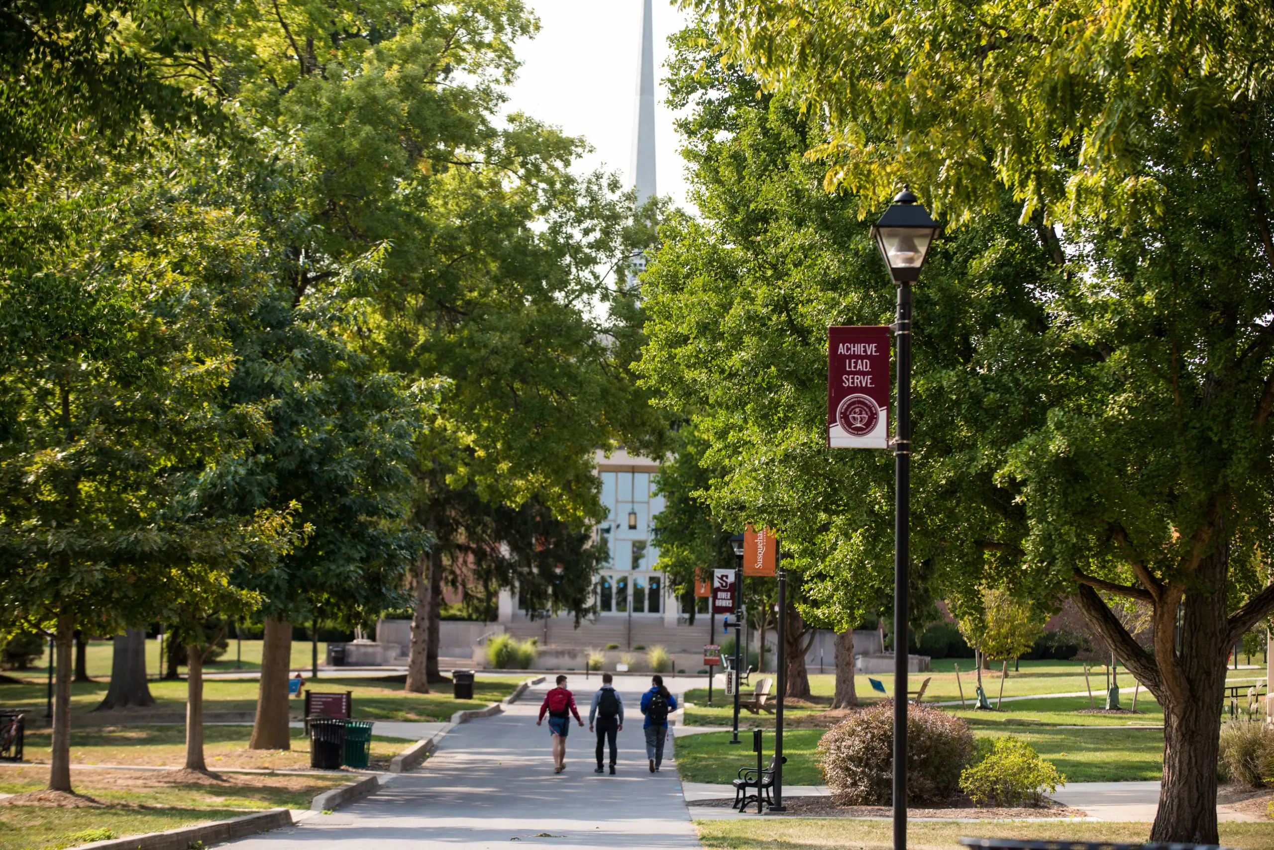 A tree-lined pathway on a college campus, with three people walking in the distance. Banners on lamp posts read "ACHIEVE LEAD SERVE." A large building with a prominent vertical structure in the background.