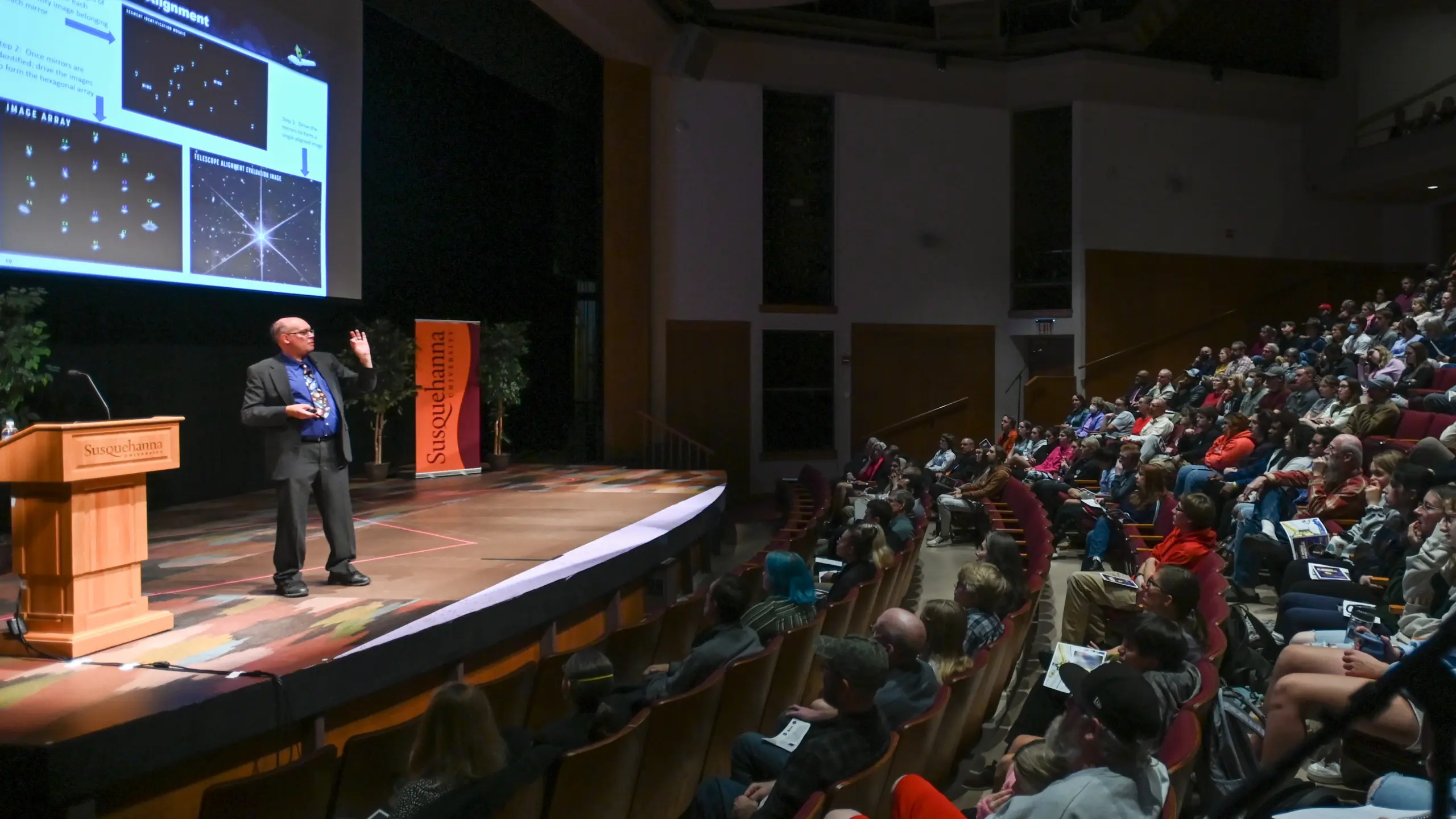 At the Claritas Distinguished Lecture in Sciences, the speaker captivates the auditorium audience with an engaging presentation. As slides illuminate the screen behind them, attendees listen attentively under the prominent "Skeptoid" banner displayed on stage.