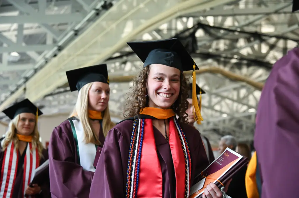 A graduate in a maroon cap and gown, with a gold tassel and honor cords, smiles and holds a program while standing in a line. Other graduates in similar attire are visible in the background, under a large, light-filled atrium.