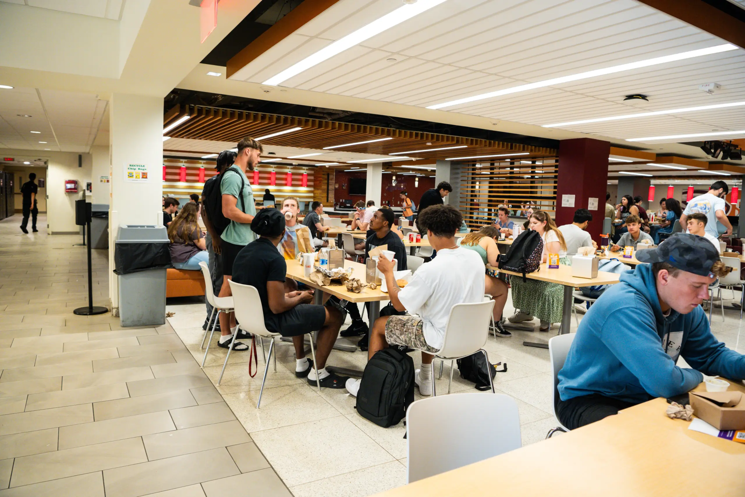 A bustling cafeteria scene with diverse groups conversing and dining, amidst the buzz of a Residence Hall Move Out day. The bright room features contemporary design elements as someone heads toward the exit, perhaps carrying boxes from their dorm room.