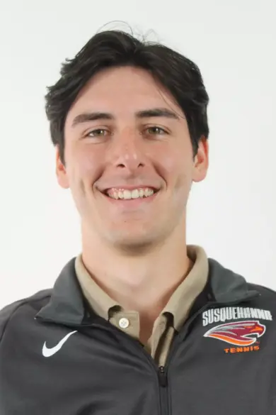 A person with short, dark hair smiles at the camera. They are wearing a jacket with a "Susquehanna Tennis" logo and a Nike swoosh. The background is plain white.