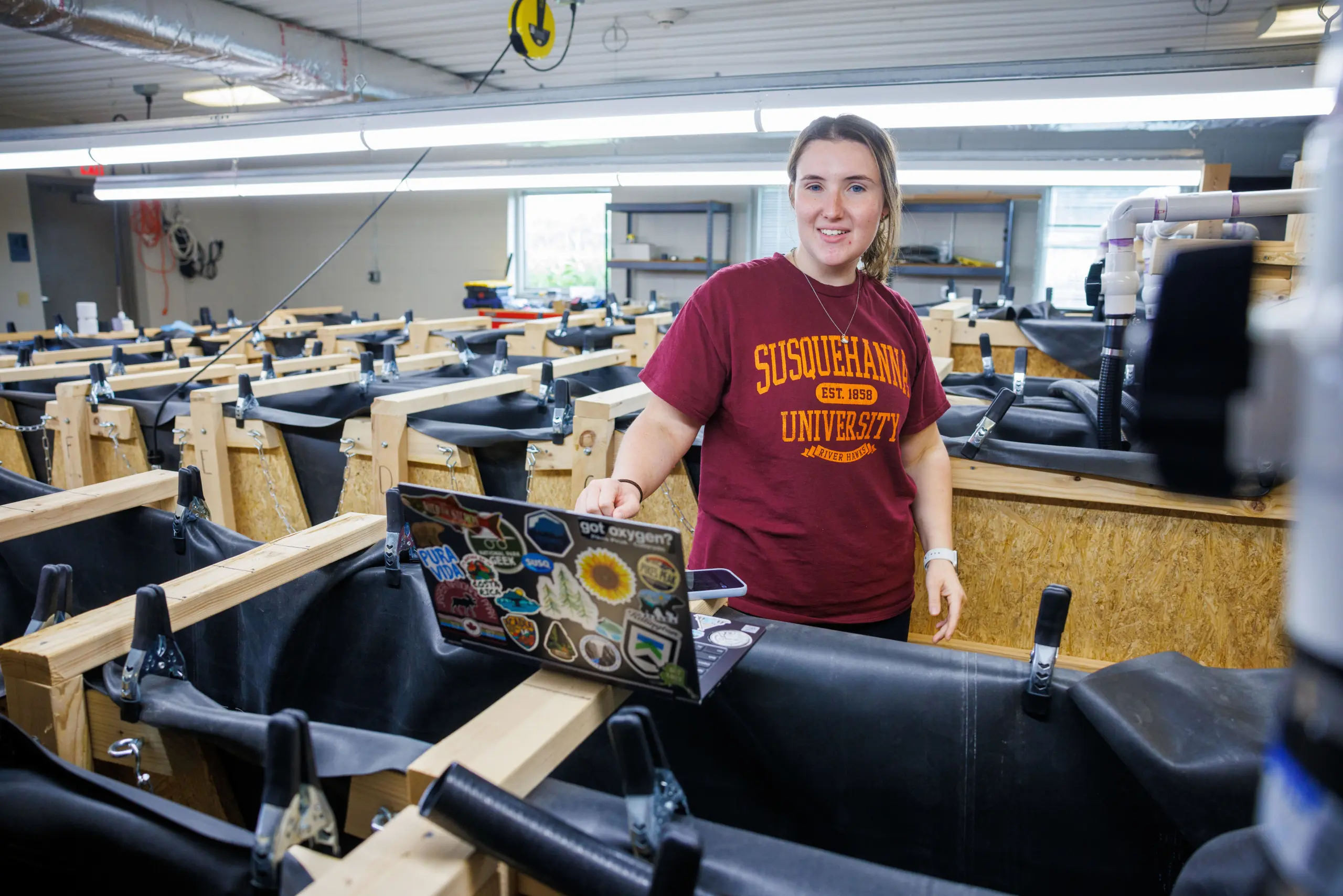 A person wearing a Susquehanna University T-shirt stands in a room with multiple tanks. They are holding a laptop covered with various stickers. The space appears to be a lab or research facility.