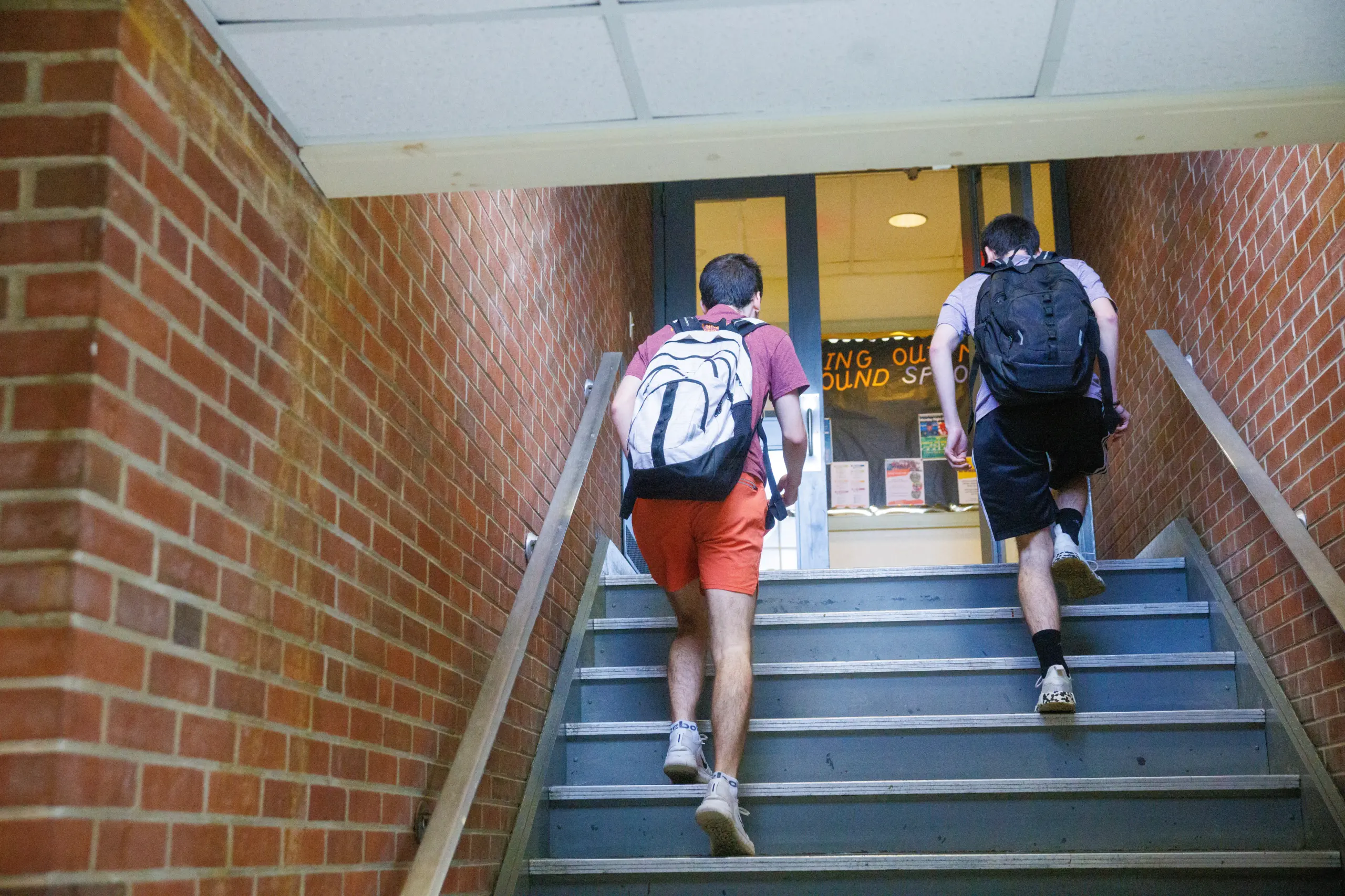 Two people with backpacks ascend a stairway in a brick building, capturing the essence of living on campus. Light spills through a windowed door at the top, where a partially visible sign hangs, creating an inviting glow in their educational journey.