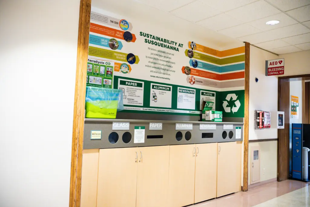 Recycling station with labeled bins for glass, paper, aluminum, newspaper, and plastic beneath a sustainability display at Susquehanna. Bright educational graphics highlight the importance of recycling and sustainability 