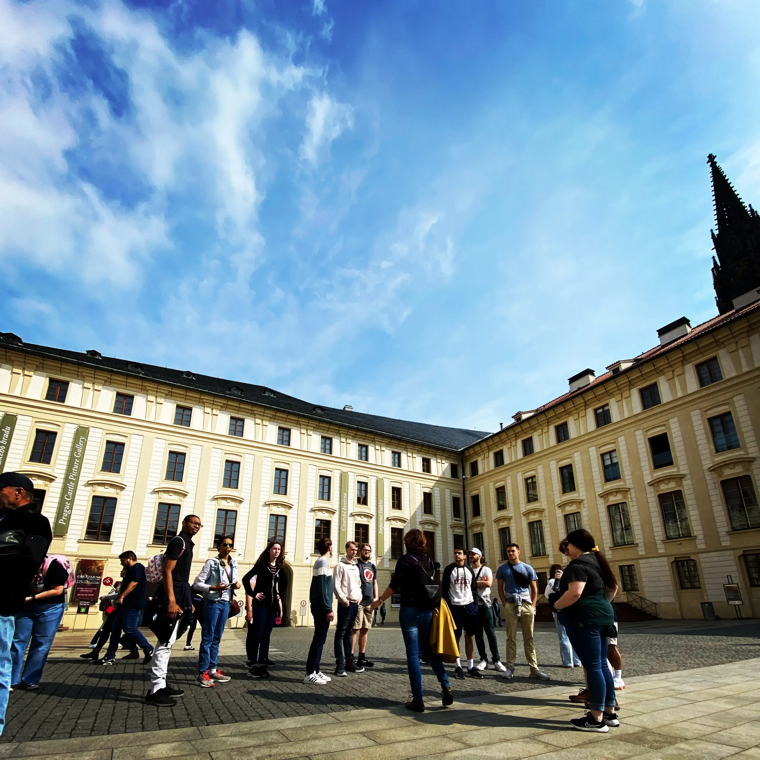 A group of tourists immersed in conversation fills a spacious courtyard surrounded by historic buildings with ornate facades under a bright blue sky, as if preparing for their next GO Study Abroad adventure.