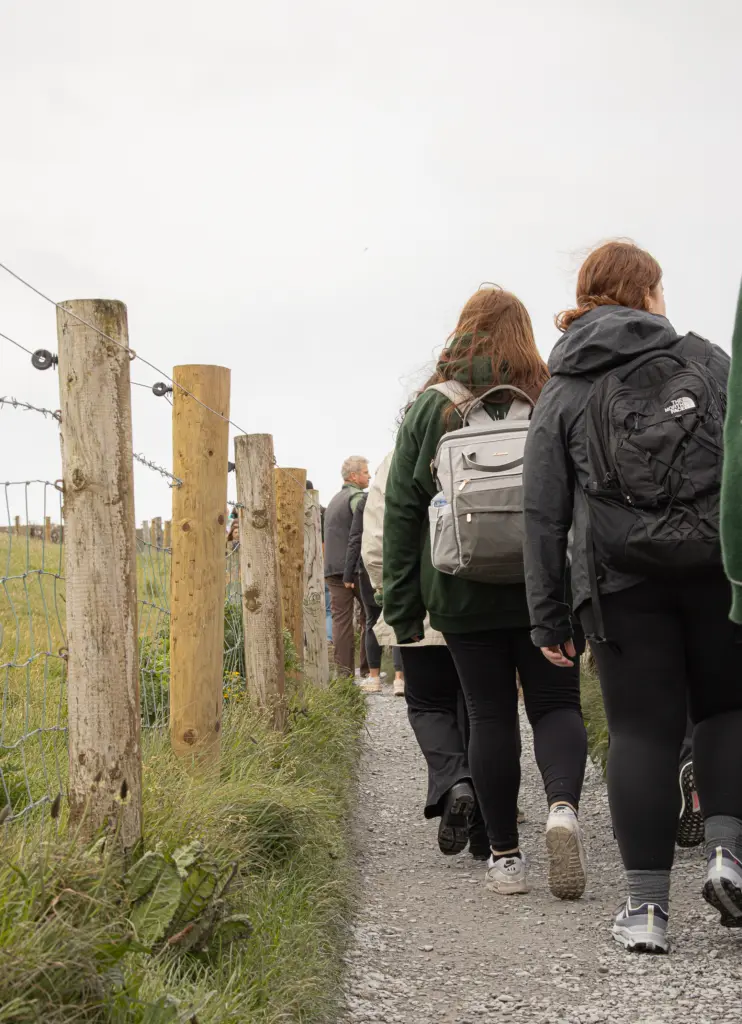 A group of people, perhaps on a GO Study Abroad adventure, walks along a gravel path bordered by wooden posts and wire fencing. Dressed in casual clothing and carrying backpacks, they wander through rural grassy fields under a cloudy sky.