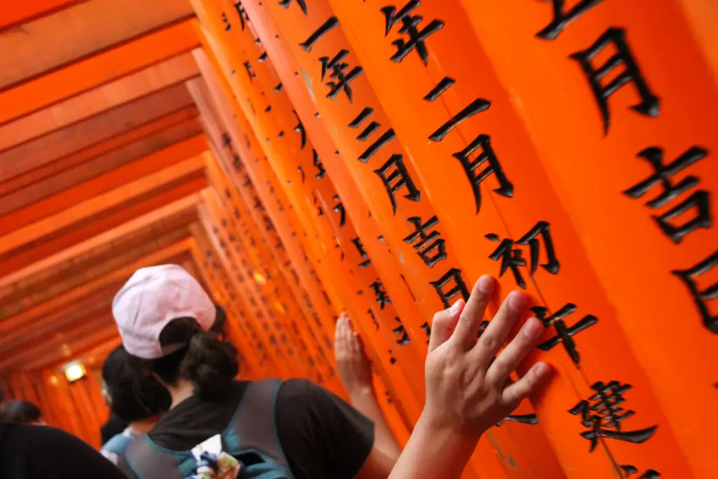 People walk through a corridor of vibrant orange torii gates with black Japanese characters at Fushimi Inari Shrine in Kyoto. A hand touches the gate, appreciating its texture and cultural significance—a moment that inspires visitors to go long on their journey of discovery.