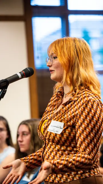 A person with orange hair and glasses speaks into a microphone at an indoor event. They wear a patterned shirt and have a name tag. Several people are seated in the background, listening attentively.