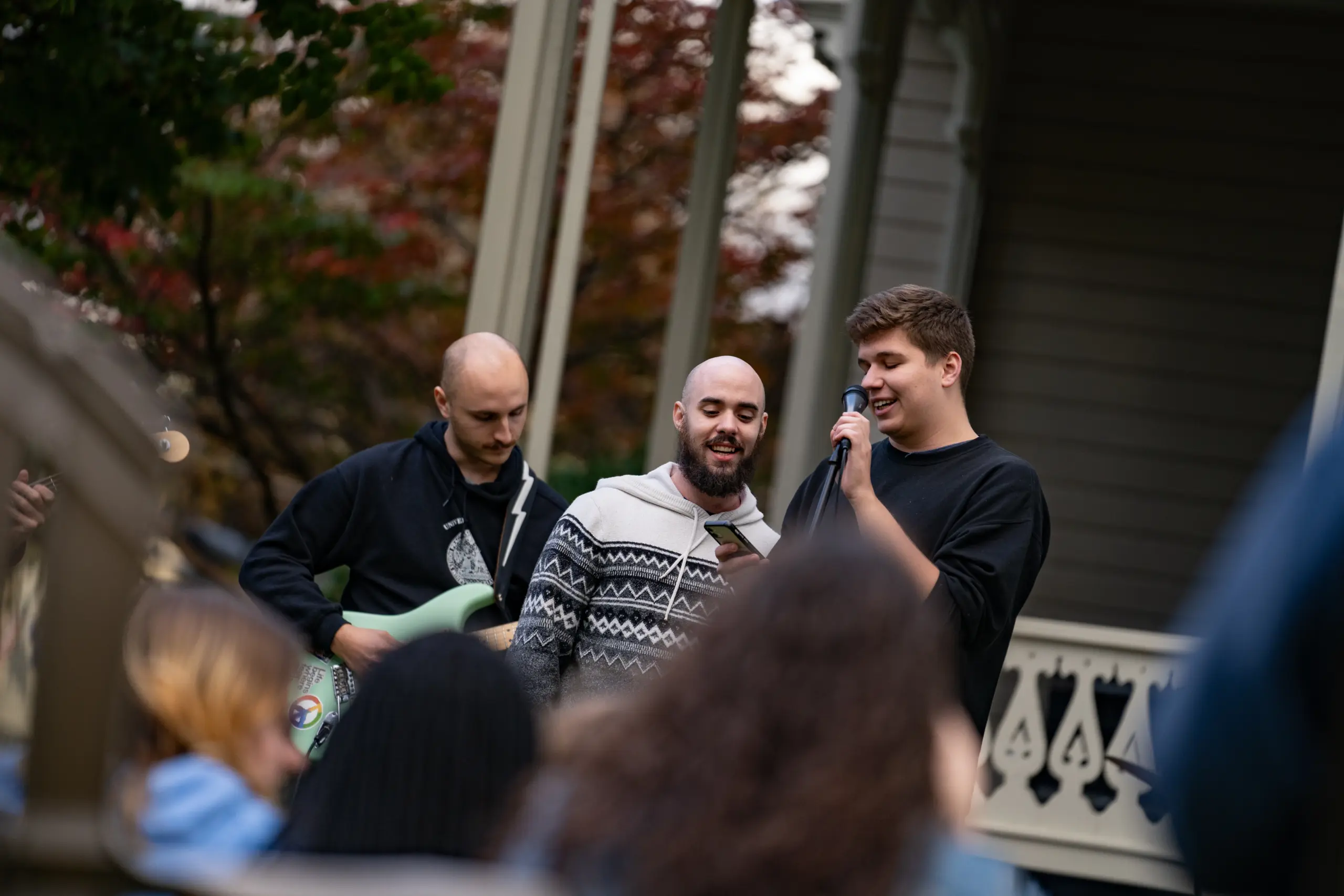 Three men are performing outdoors on a small stage, adding a harmonious vibe akin to Greek life. One strums a guitar while the other two sing into microphones. Casually dressed, they draw a small crowd, with a porch and autumn trees providing the perfect backdrop.