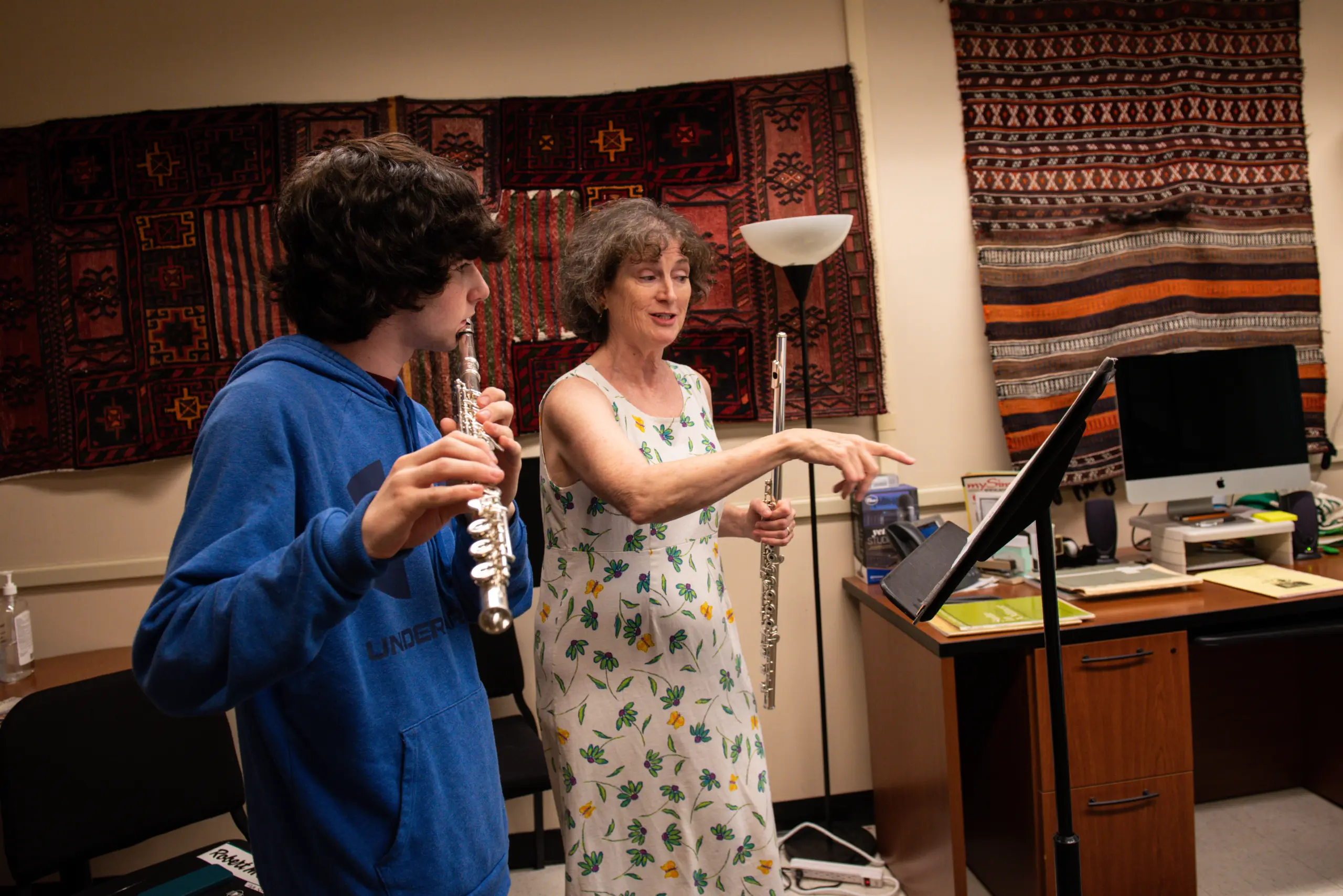 A person in a blue hoodie plays the flute, receiving guidance from a woman in a floral dress holding a flute. They're in a room with colorful tapestries on the wall, a floor lamp, and a music stand with sheet music between them.