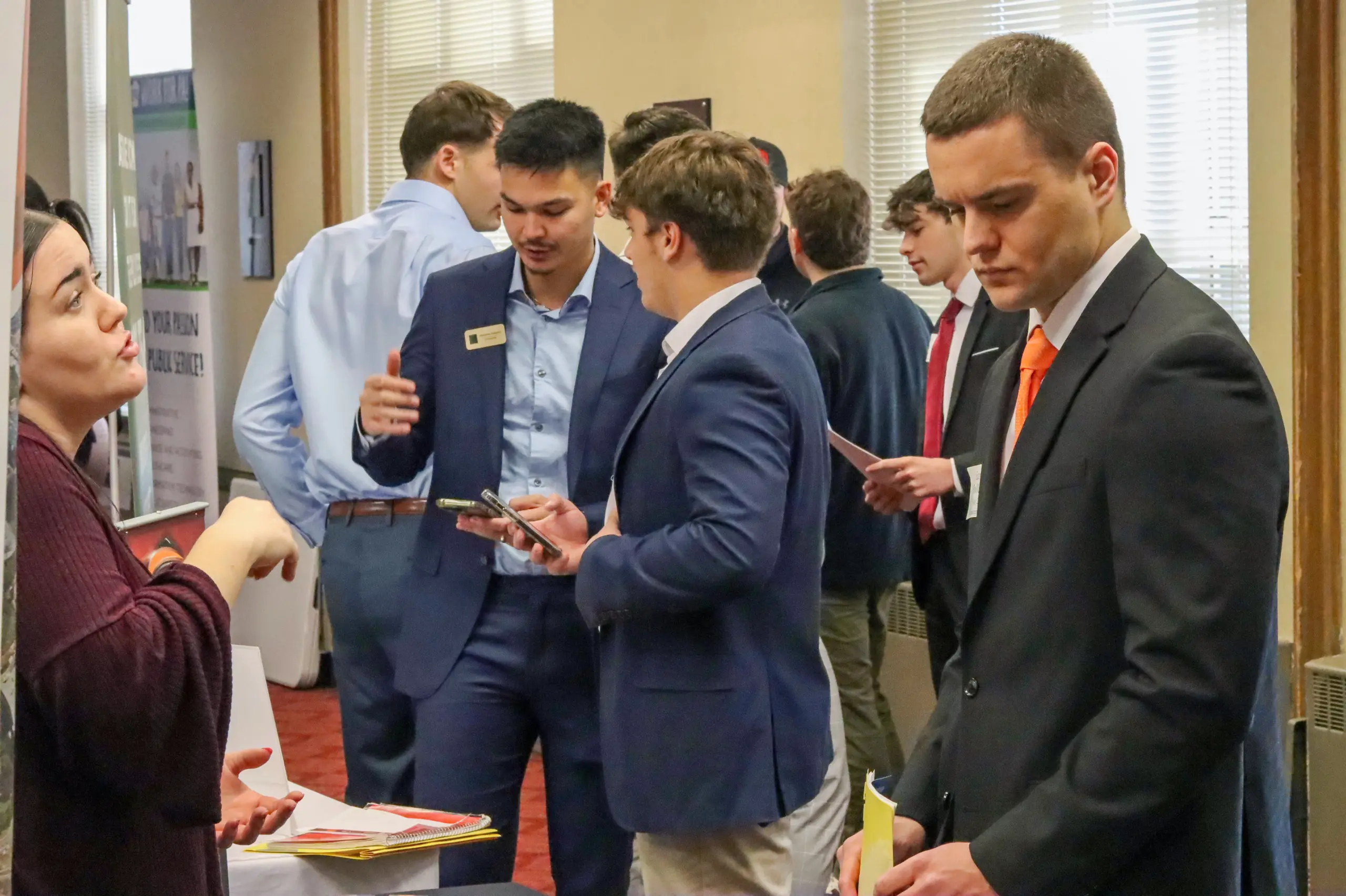 A group of people in business attire engage in conversation at a professional networking event. Some are holding papers and phones. A woman is gesturing while talking to a man. The setting is an indoor conference or meeting room.