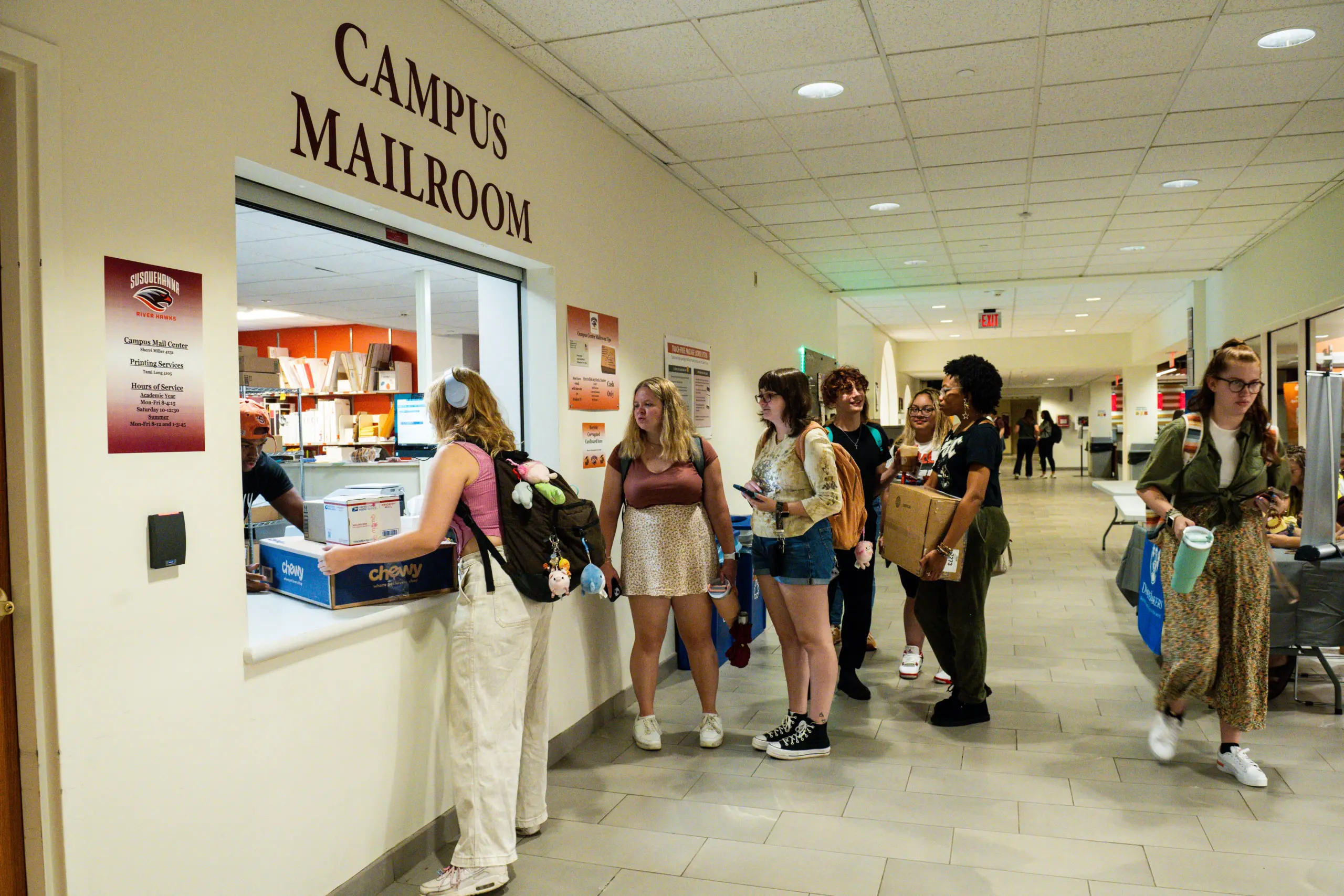 Students line up at a campus mailroom window. One person at the counter hands over a package, while others wait holding boxes and papers. The setting is a brightly lit hallway with office doors and bulletin boards visible.