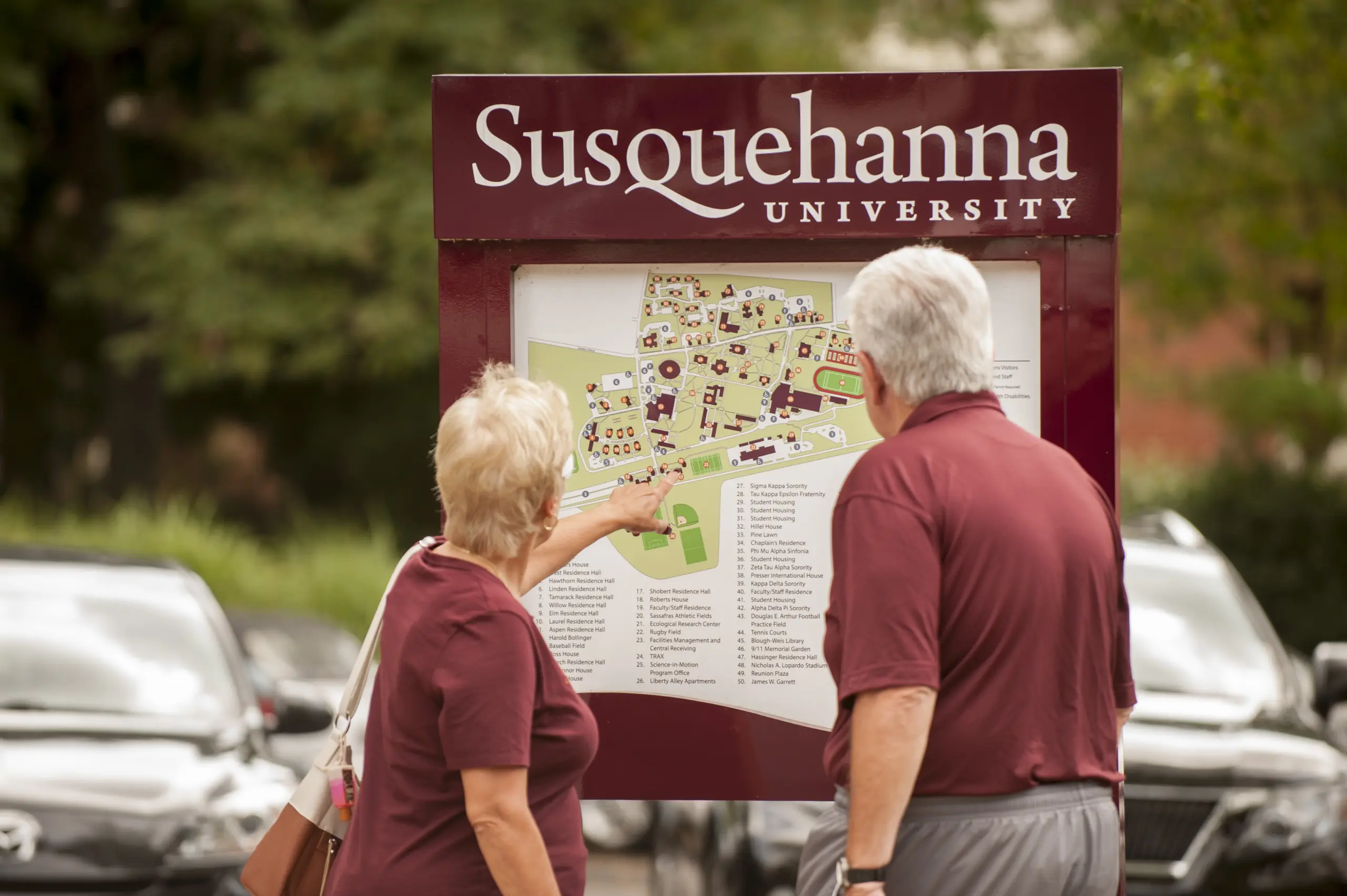Two people in maroon shirts consult the Maps and Directions sign labeled "Susquehanna University." The woman points to a spot on the campus map. They stand amid parked cars with lush greenery in the background.