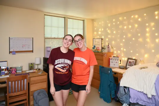 Two people stand smiling in a cozy first-year housing dorm room. Both wear Susquehanna University T-shirts, one in maroon and the other in orange. The room features a neatly organized desk, a bed, twinkling string lights, and various decorations.