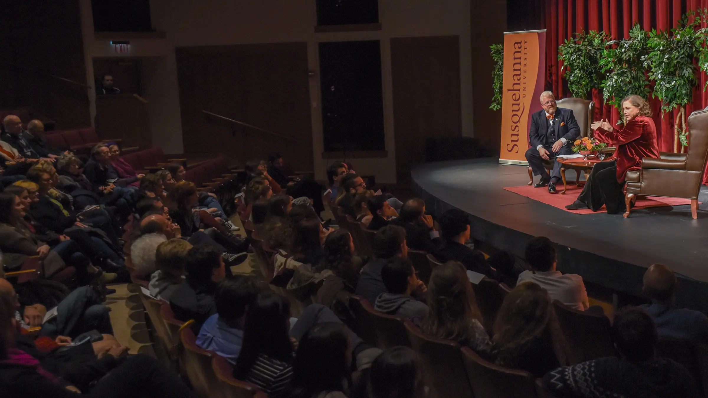 Two people are seated on stage in a dimly lit auditorium for the Dr. Bruce L. Nary Theatre Guest Artist Lecture. A red curtain and a "Susquehanna" banner hang in the background as an attentive audience fills the theater seats, absorbing every word of their engaging discussion.