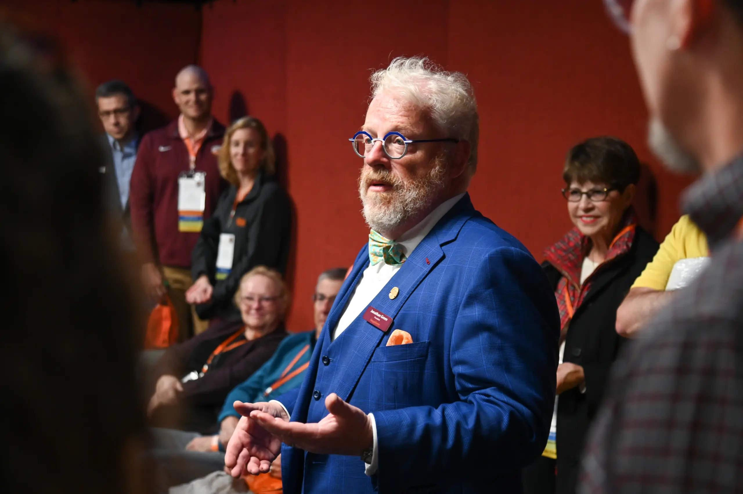 A person wearing a blue suit and bow tie speaks animatedly to a group in a room with red walls. Several people, some seated and others standing, listen attentively.