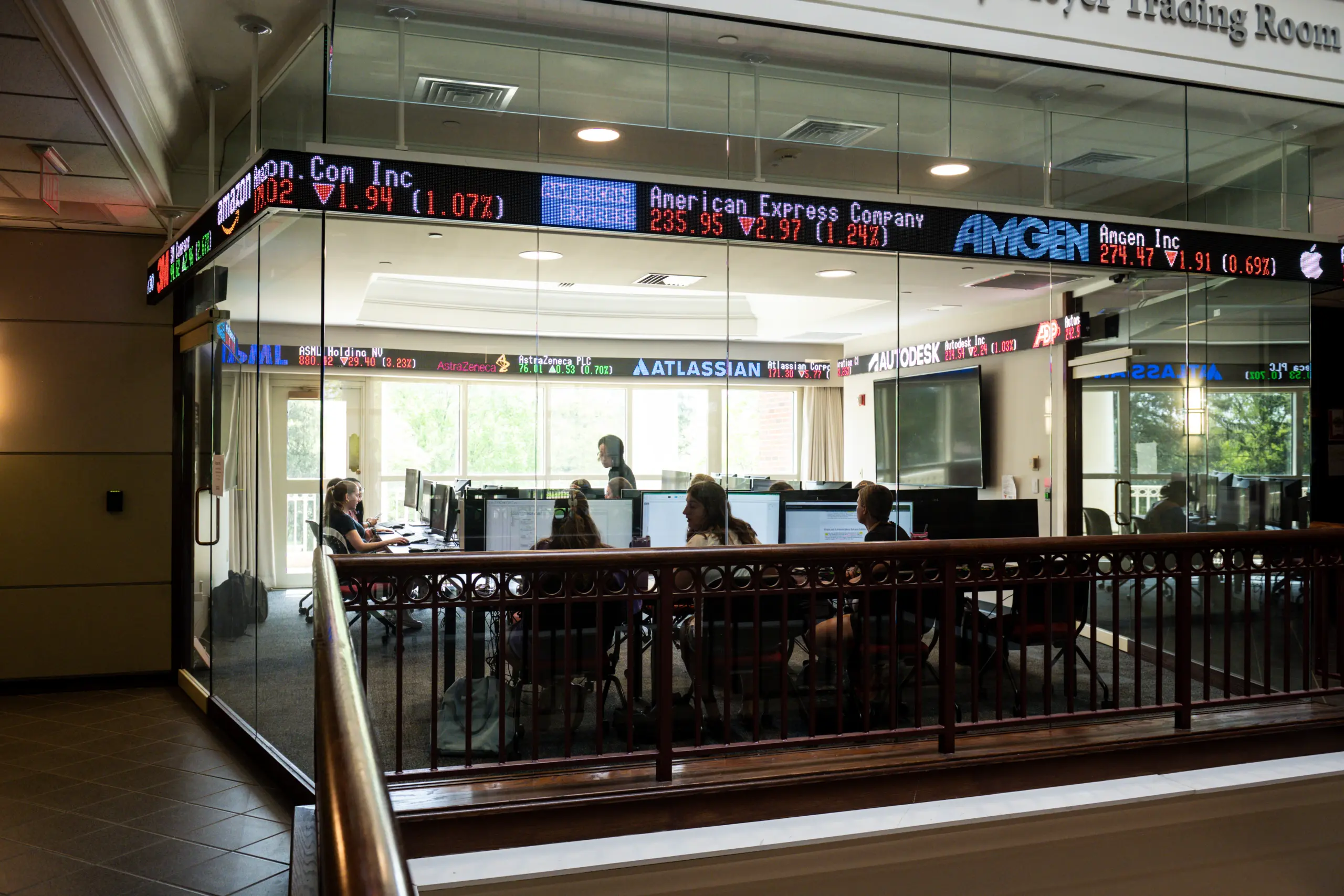 A glass-walled classroom with students seated at desks, computers, and stock tickers displaying company names like AMGEN and American Express. Natural light streams through large windows, creating a focused and modern atmosphere.