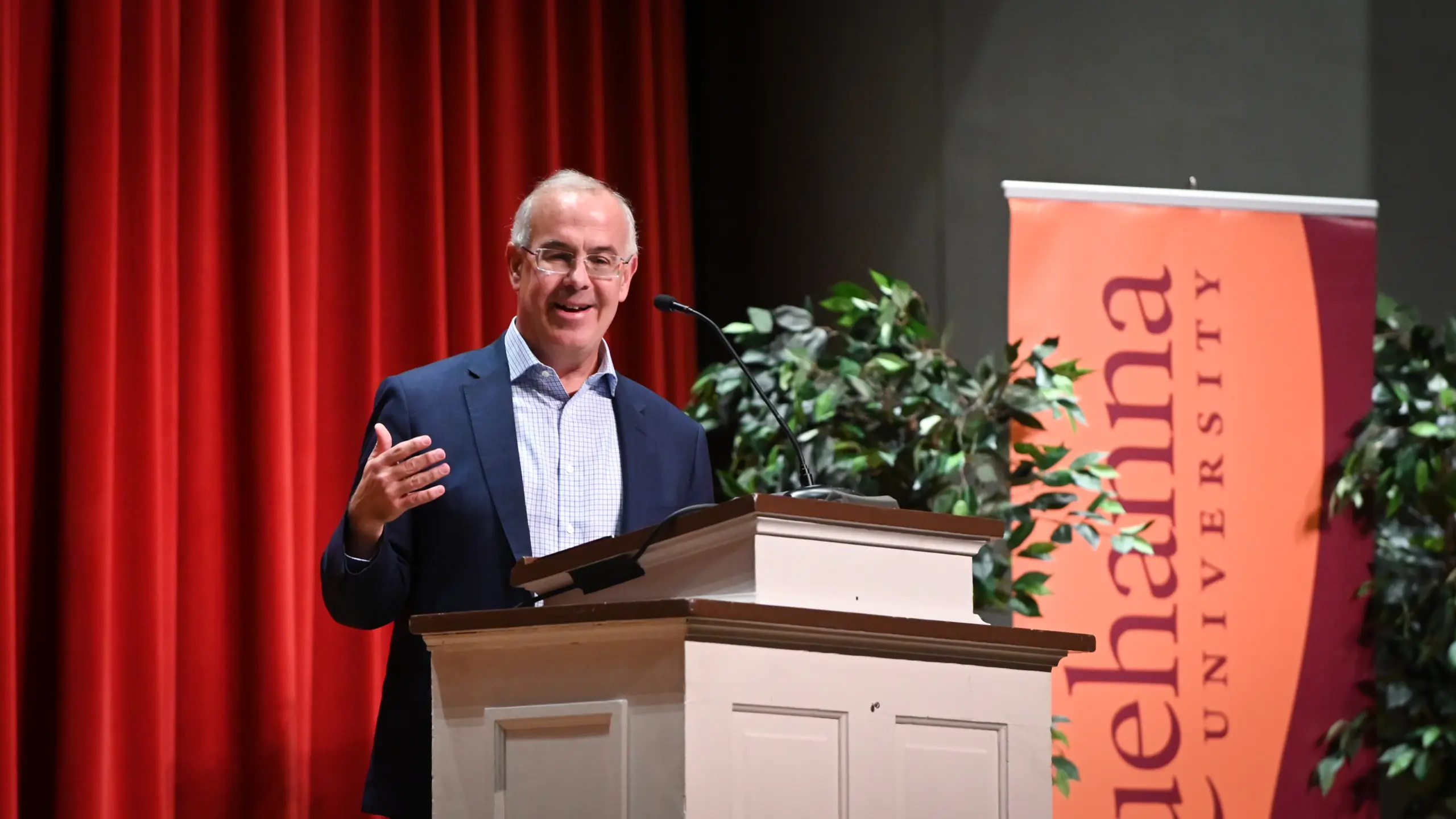 A man stands at a podium, gesturing with one hand while speaking during the Edward S. & A. Rita Schmidt Lectureship in Ethics. He is wearing a suit and glasses. Behind him hangs a red curtain and a banner displaying the partial words "hanna University.