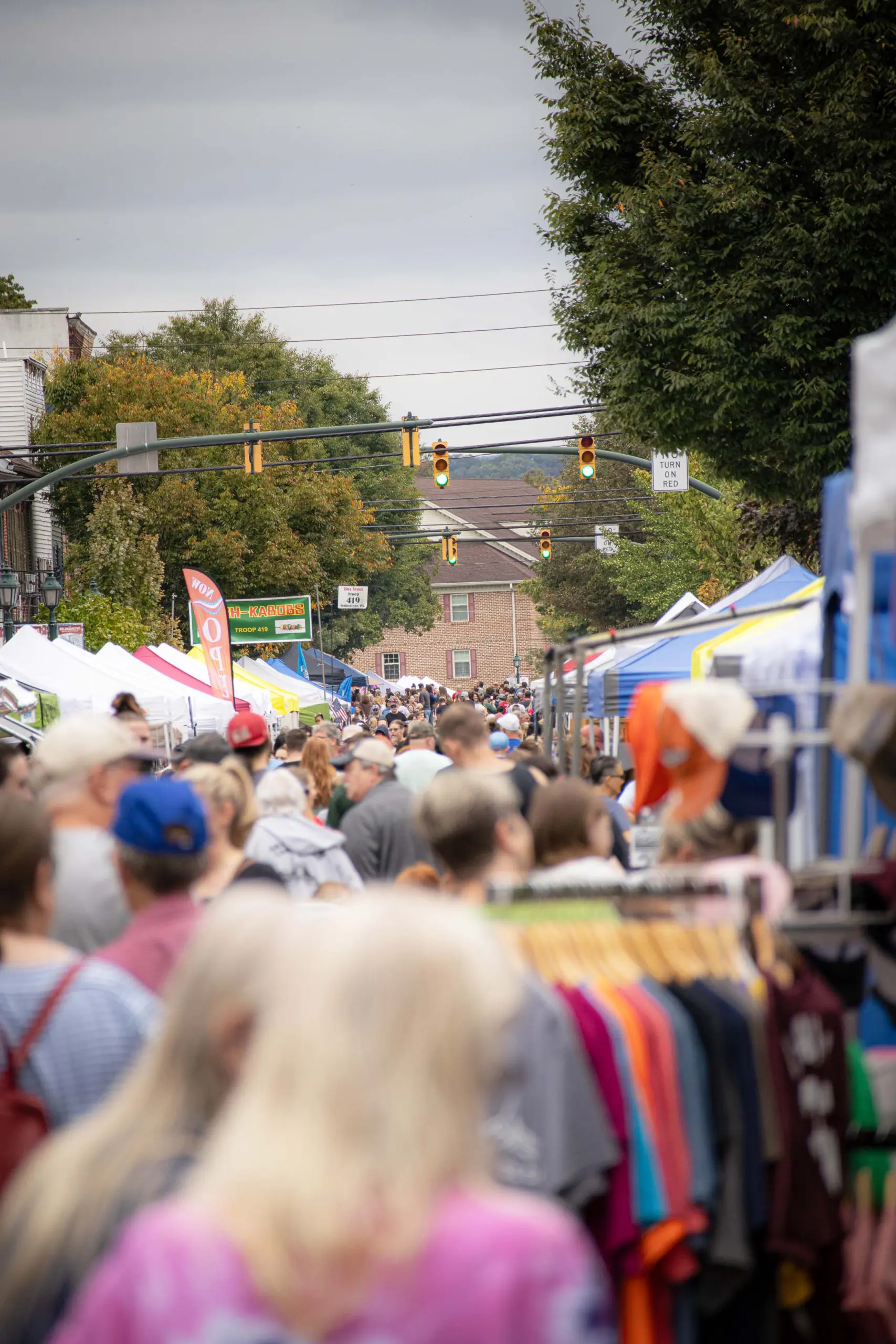 An about bustling outdoor market scene unfolds on a street dotted with numerous white and blue tents. People stroll through, browsing various stalls selling clothing and other items. The overcast sky looms above while green trees line the background, adding to the vibrant atmosphere.