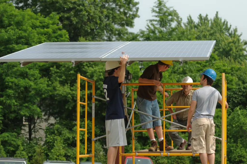 Four people are installing solar panels on a metal frame in an outdoor setting. They are wearing hard hats and casual clothing. Lush green trees are visible in the background.