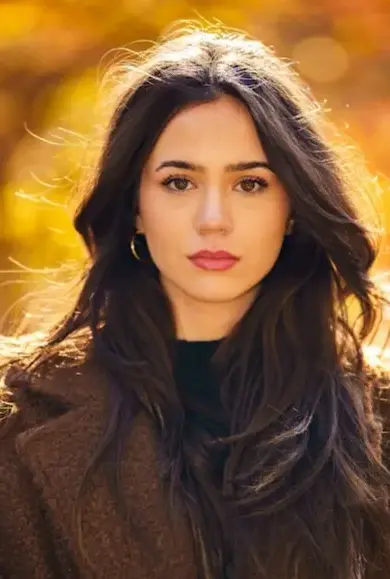 A woman with long dark hair stands in a sunlit outdoor setting with a blurred background of autumn leaves. She wears a brown coat and a serious expression, looking directly at the camera.