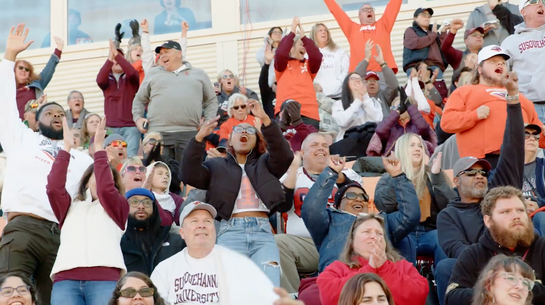 A diverse crowd at a sports event, wearing various team apparel, cheering enthusiastically with raised arms. The scene captures a mix of emotions, including joy and excitement, as spectators engage with the game.