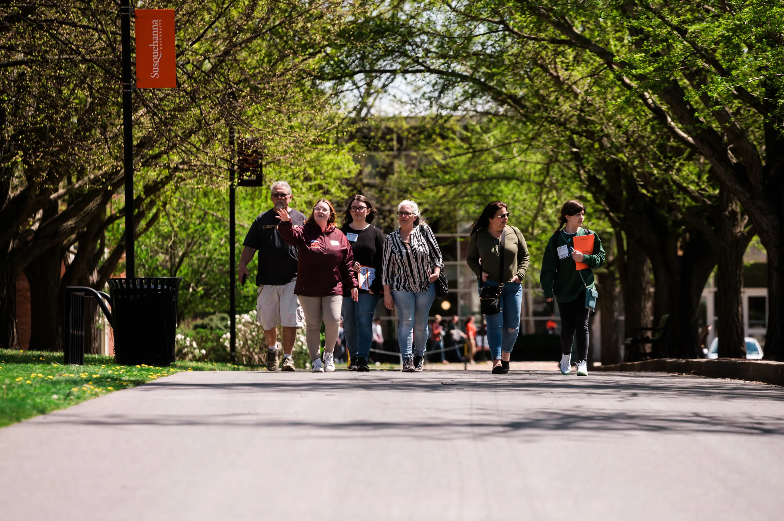 A group of six people, casually dressed, walk along a tree-lined campus path. They seem engaged in conversation. The season appears to be spring with trees in full leaf. A banner with text is visible in the background.
