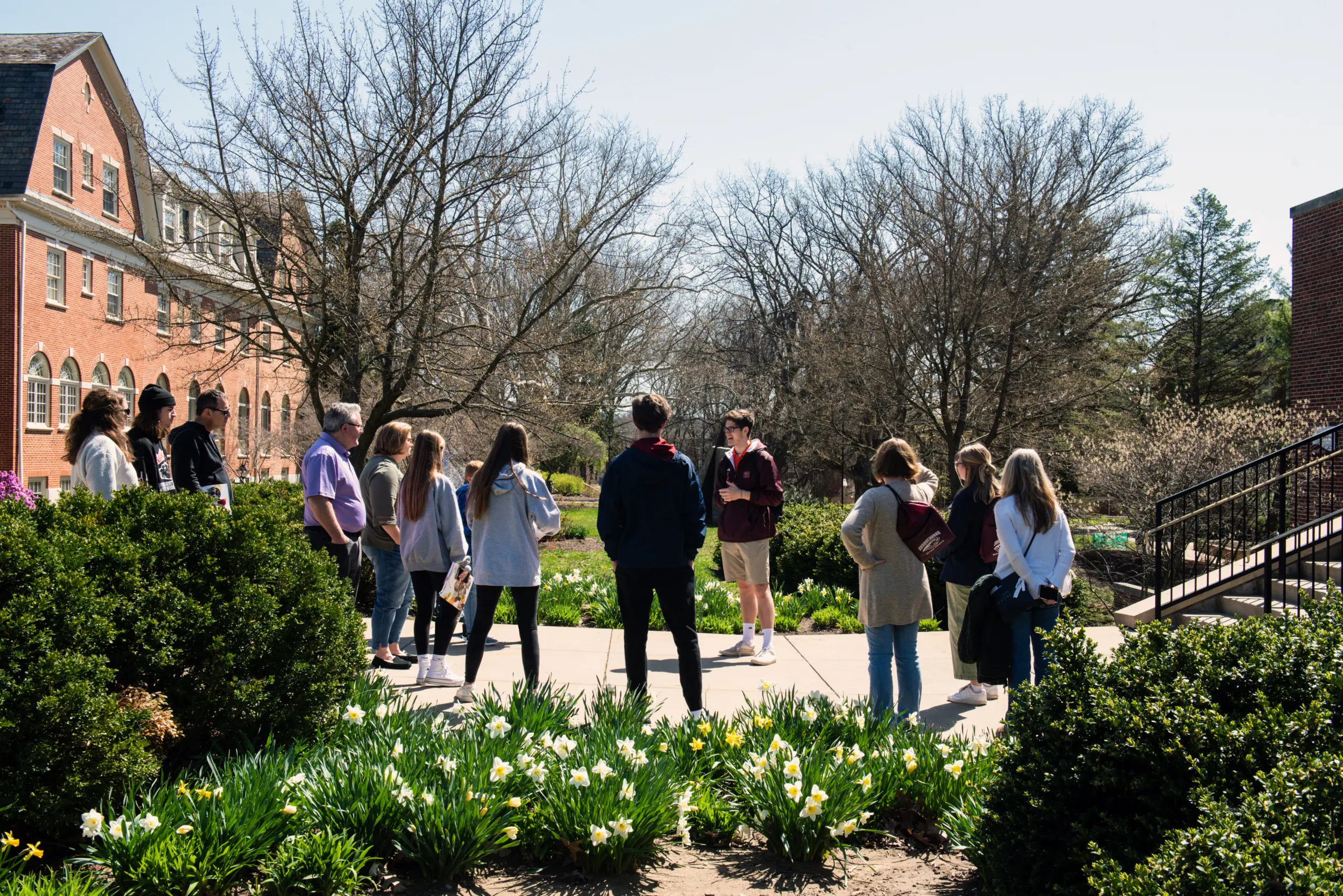A group of people stands on a paved path, listening to a person speaking in front of them. The scene is outdoors, with trees without leaves and a brick building in the background. Green bushes and blooming flowers are in the foreground.
