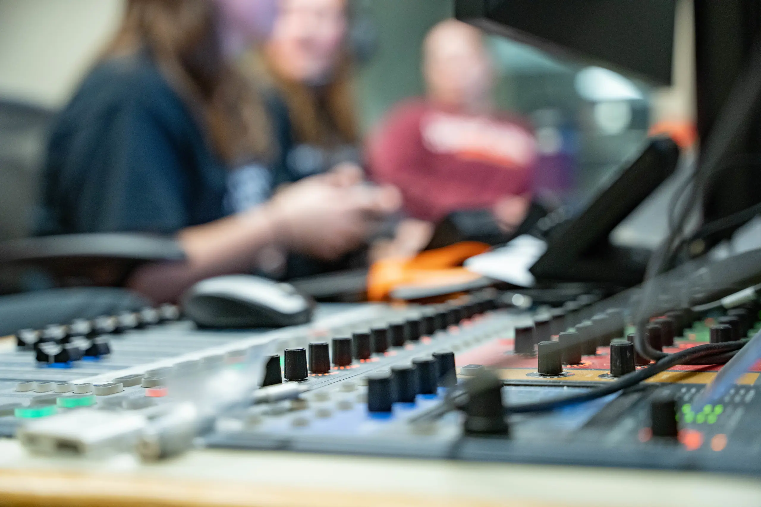 Close-up of a mixing console with various switches and knobs in focus, embodying a "Learn By Doing" approach. In the blurred background, three people are engaged in conversation or work, with a computer monitor partly visible on the right, illustrating hands-on collaboration.