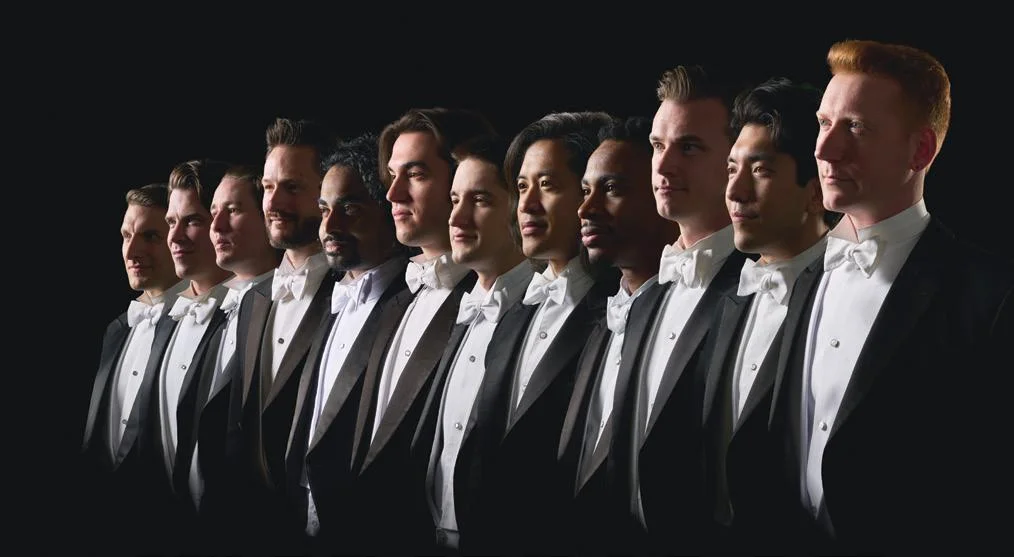 A group of twelve men in formal attire, wearing black tuxedos with white bow ties, standing side by side in a line against a dark background. They are all looking confidently forward.