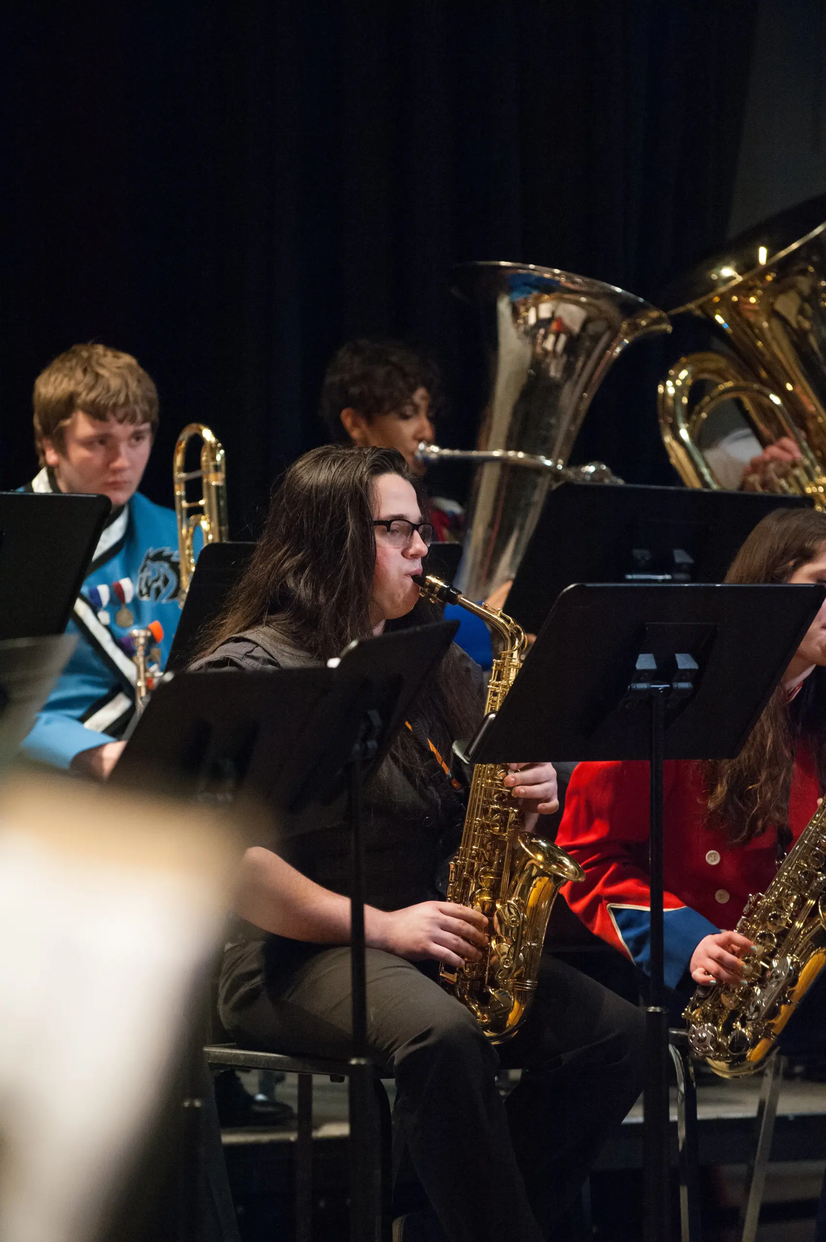 A band performing at a music festival features a saxophonist in the foreground wearing a black shirt. In the background, musicians play brass instruments like tubas, with music stands set before them, creating an electrifying workshop of sound and rhythm.
