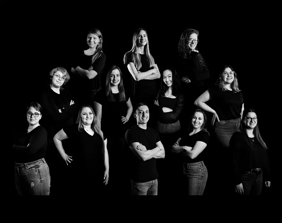 A black and white group portrait, reminiscent of an art piece from the Lore Degenstein Gallery, features twelve people posed against a black background. Arranged in two rows, all wear dark clothing. Most are smiling, with arms crossed or on hips, exuding confidence.