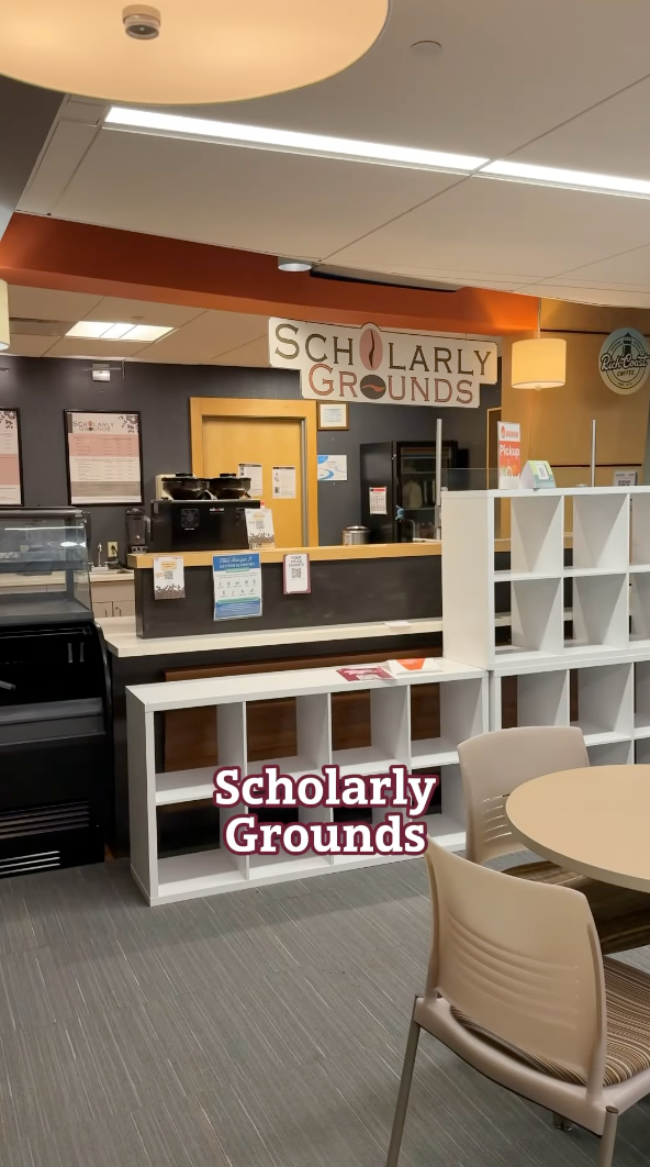 Interior view of "Scholarly Grounds," a campus haven where academia meets modern design. Cozy shelves line the walls, and students gather around a round table, sipping espresso. The menu above the counter boasts scholarly-themed brews—a perfect spot for living on campus with vibrant intellectual energy.