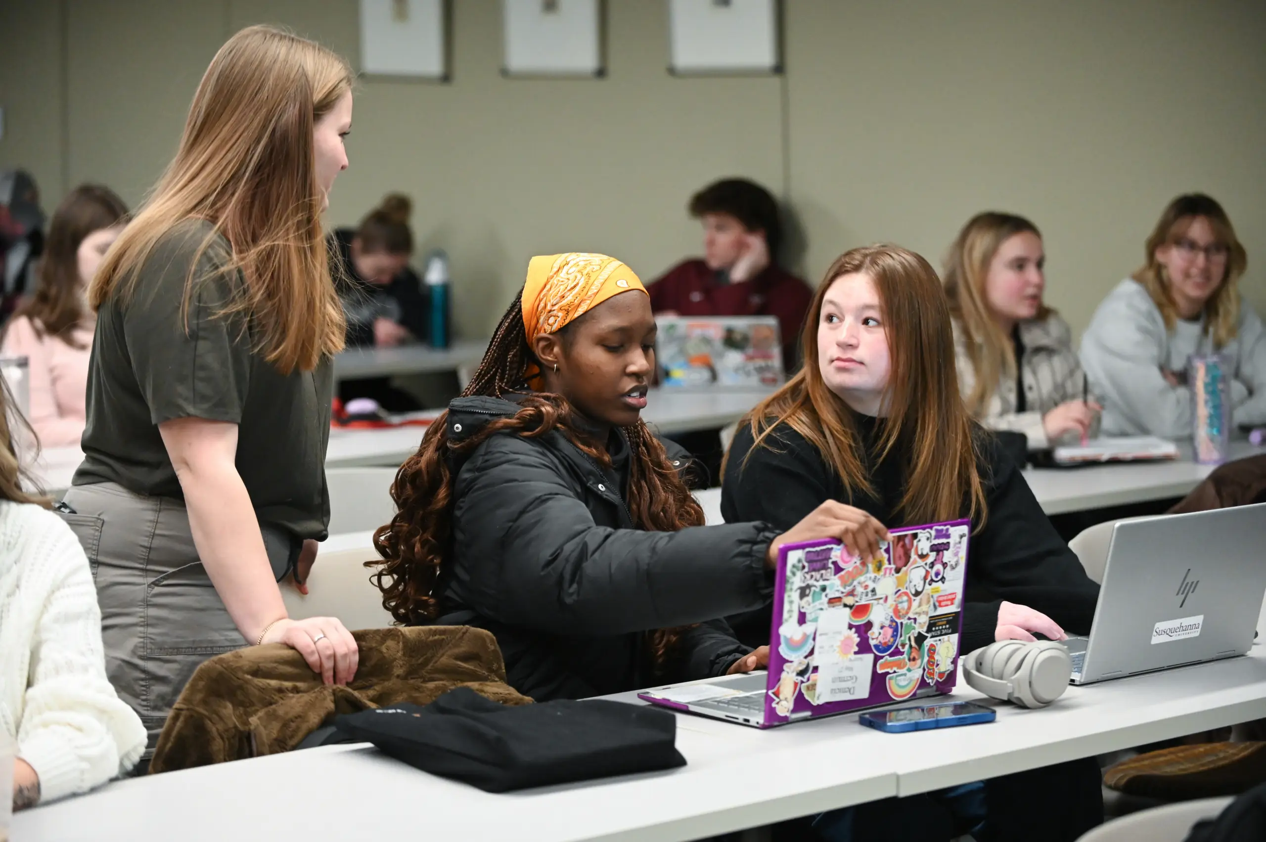 Students in a classroom are engaged in conversation. A woman standing chats with two seated women, one with a decorated laptop. Other students are visible in the background, some paying attention, others focused on their laptops.