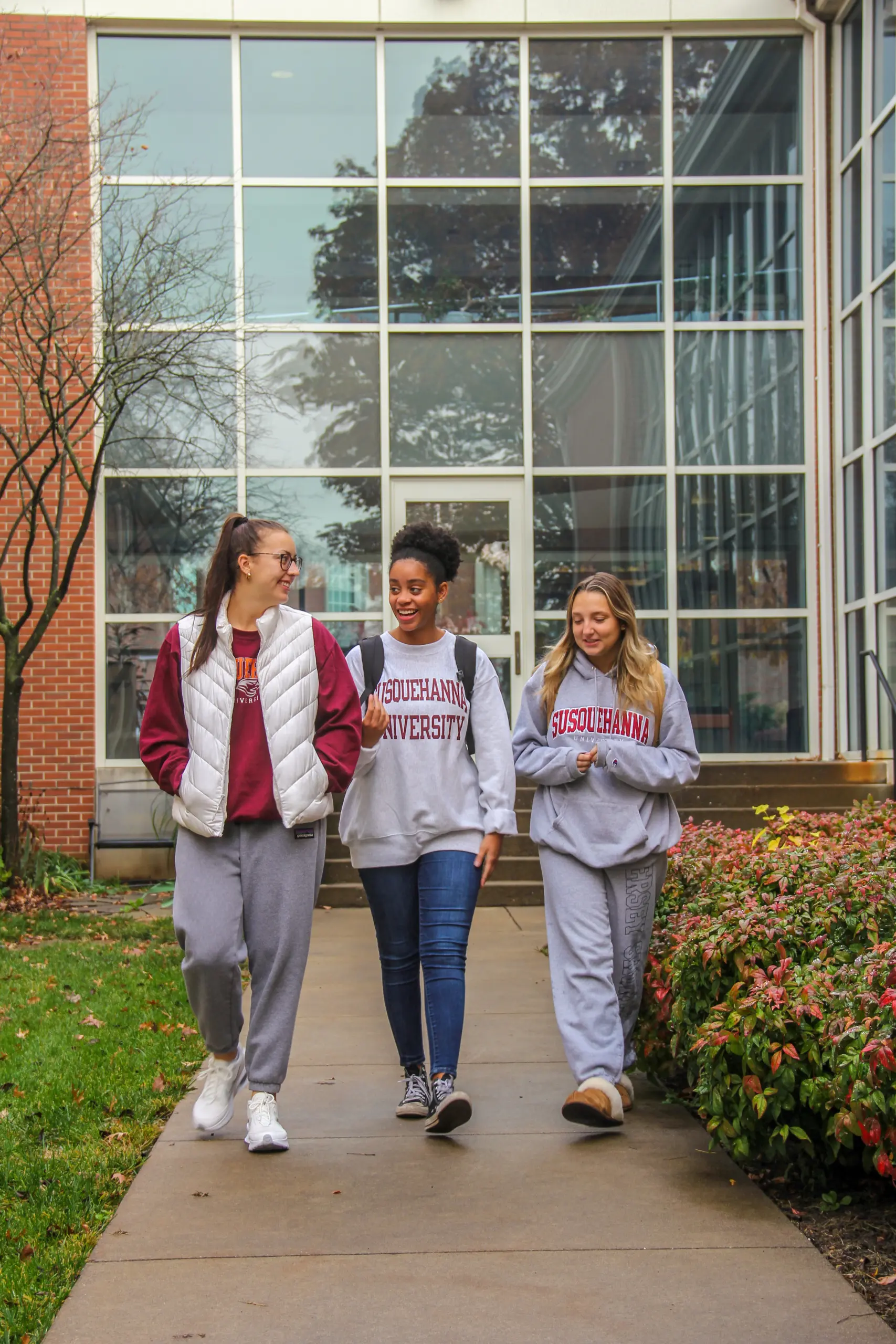 Three students walk together outside a building with large windows. They wear Susquehanna University apparel and carry backpacks. The path is lined with shrubs, and a tree with bare branches is visible on the grassy area.