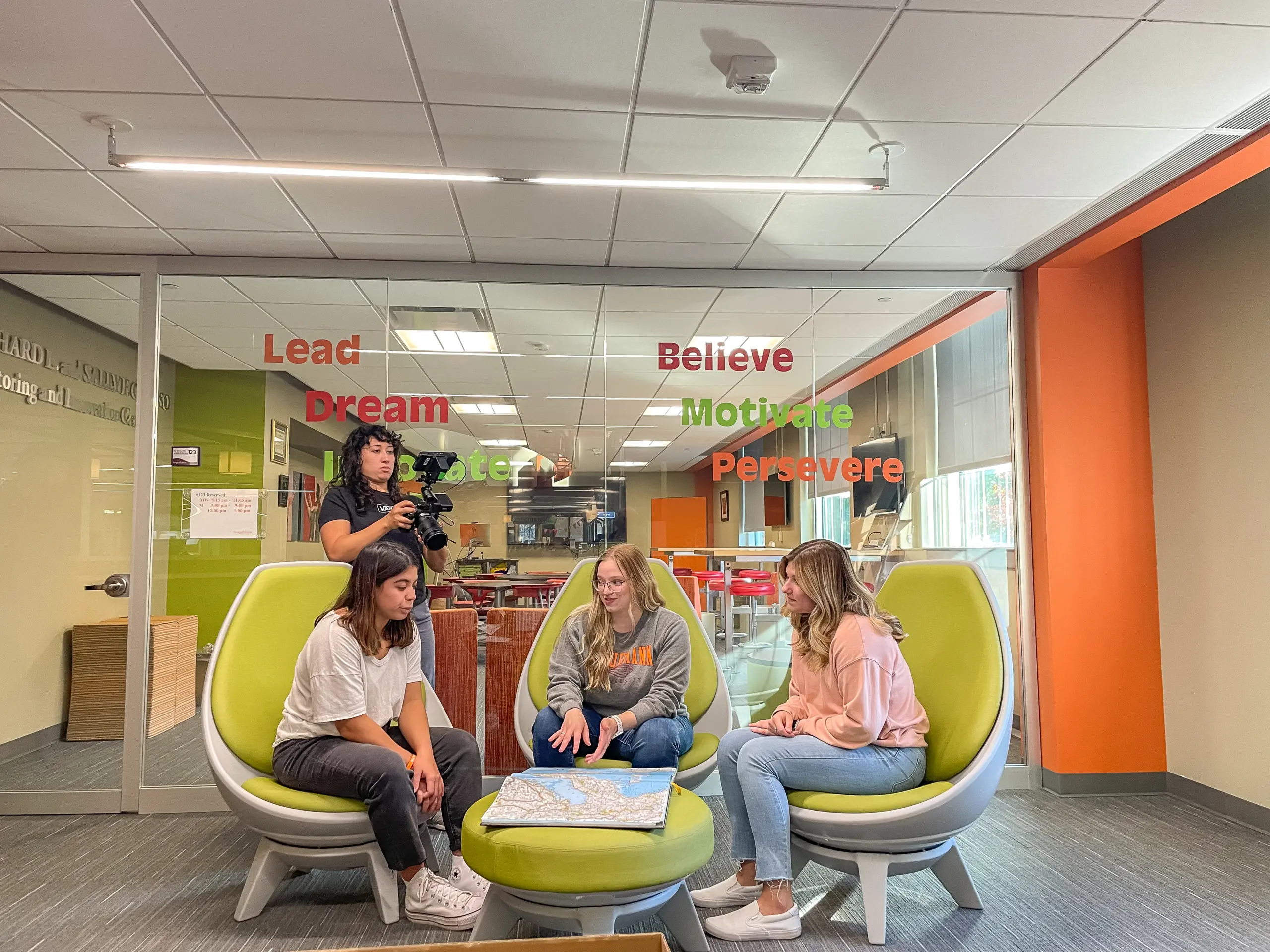 Four people sit on green chairs around a table with a map in a modern room. Wall text reads "Lead, Dream, Believe, Motivate, Persevere." One person holds a camera, while others look at the map and chat. Bright, colorful decor enhances the lively space.