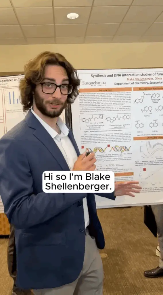 A person in a blue suit and glasses stands in front of a scientific poster at an academic conference. A speech bubble from the person reads, "Hi, I'm Blake Shellenberger." The poster is filled with detailed chemical diagrams and graphs.