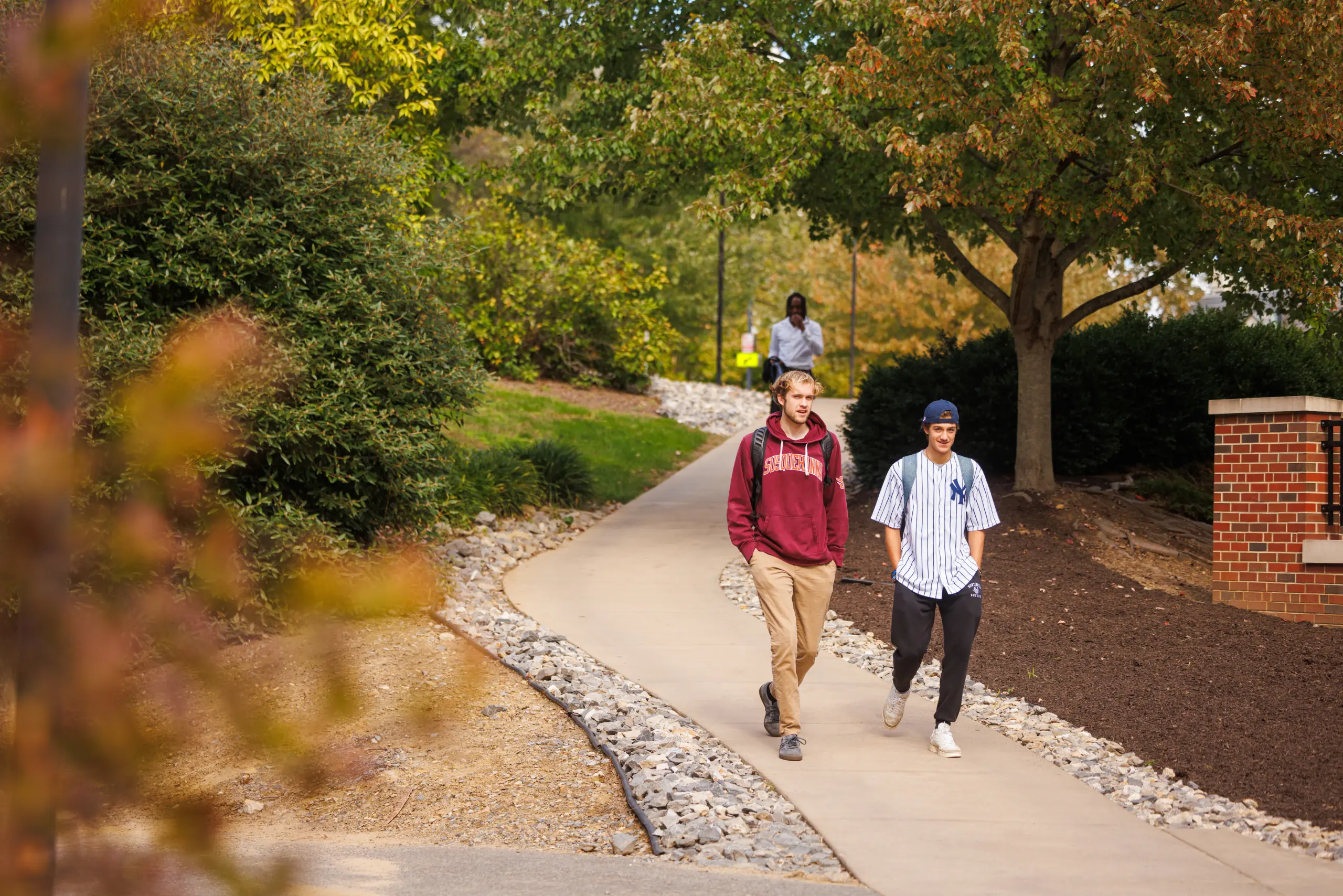 Two people walk along a paved path through a park. One wears a red hoodie and khaki pants; the other wears a striped baseball jersey and dark pants. Lush green trees surround them, and a third person is seen in the background.