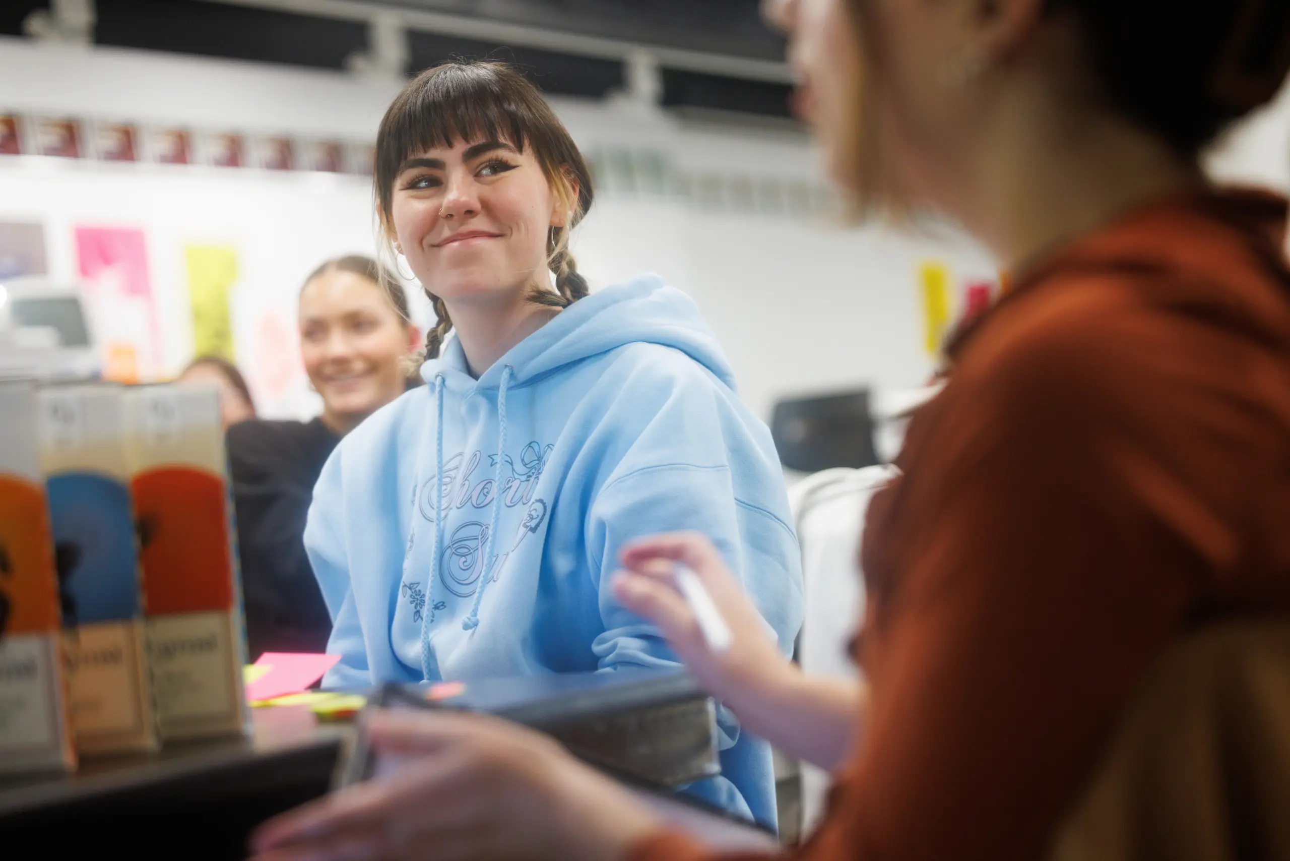 A person in a blue hoodie with braided hair smiles at someone speaking in the foreground. They are seated with others in a bright room, suggesting a casual or classroom setting.