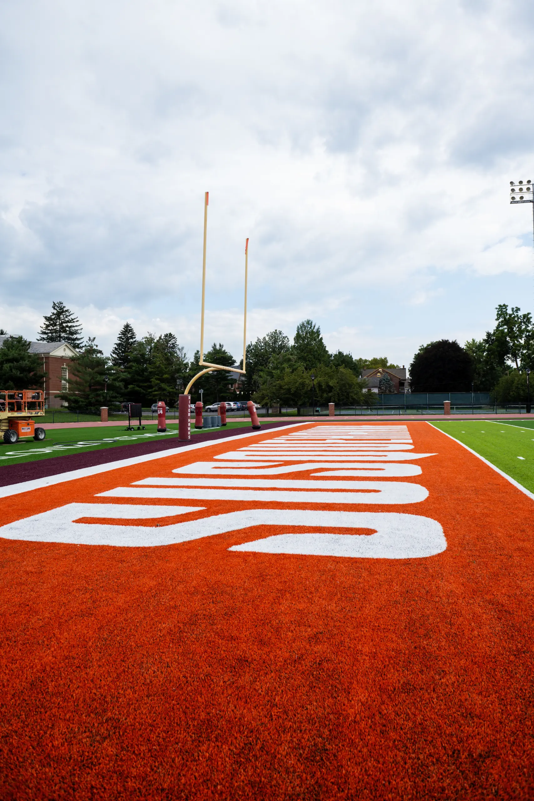 Close-up of a football field's end zone, featuring bright orange turf with the word "Indians" in large white letters, embodies the spirit of athletics and recreation. Yellow goalposts stand against a partly cloudy sky, framed by trees along the perimeter.