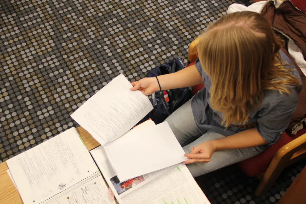 A person with long blonde hair is sitting on a chair, looking at papers on a table. The table has open notebooks and books. The person is wearing a blue shirt and jeans, with their focus on the documents in their hand.
