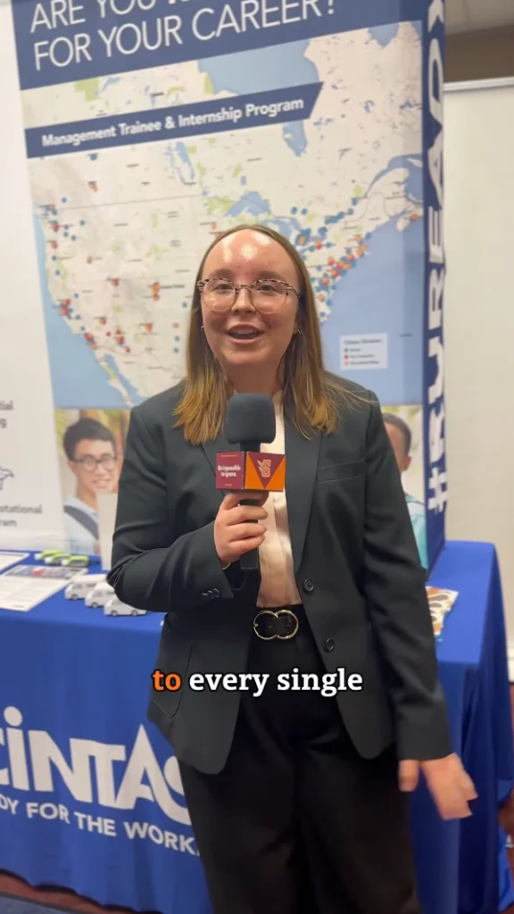 A person in a black blazer is standing with a microphone in front of a booth with a banner that says "ARE YOU READY FOR YOUR CAREER?" and a map of the United States. The table is covered with informational material and a blue tablecloth.
