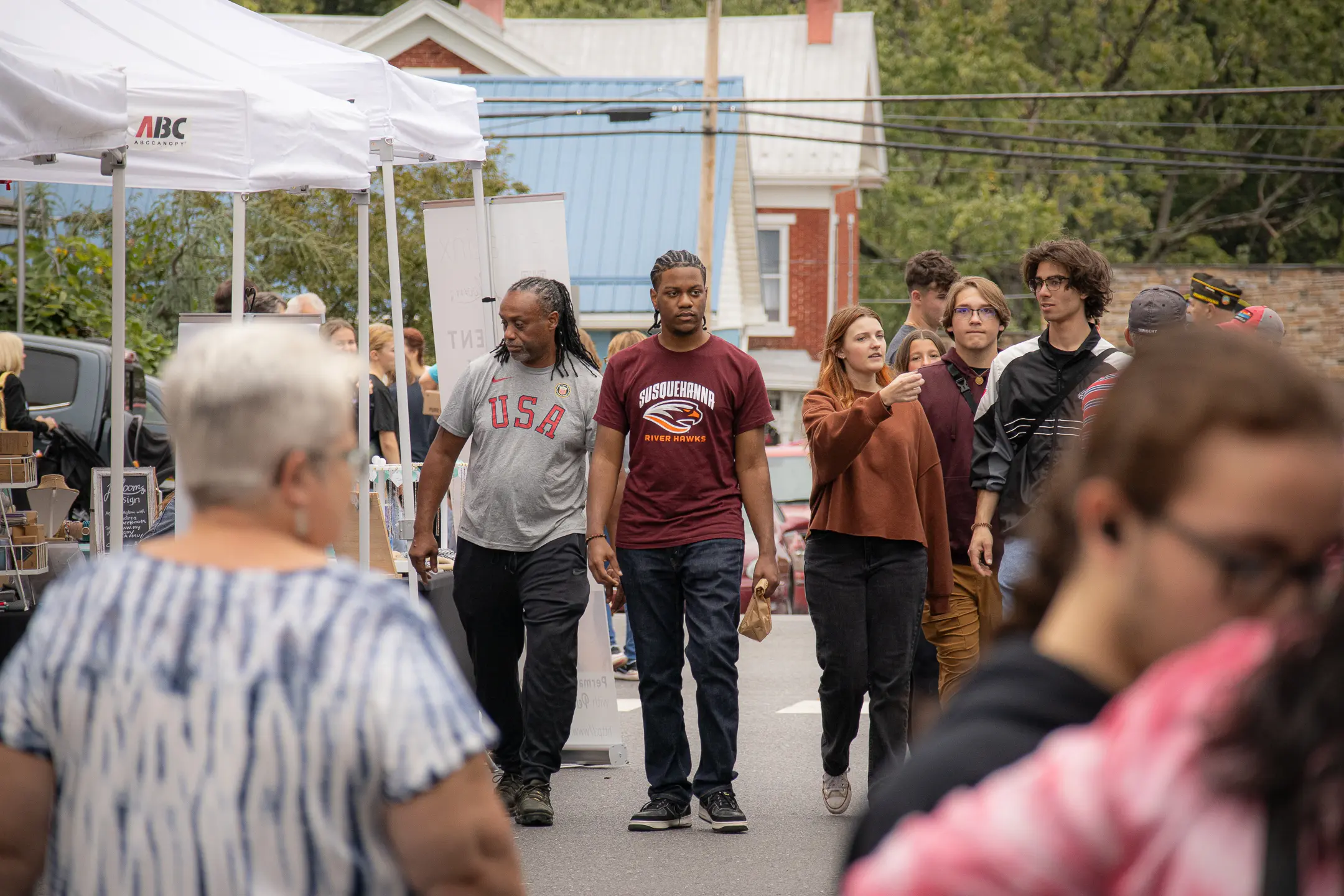 A diverse group of people walking through an outdoor market. Stalls covered with white canopies line the street. Some individuals are engaged in conversation, while others browse. Buildings and greenery are visible in the background.