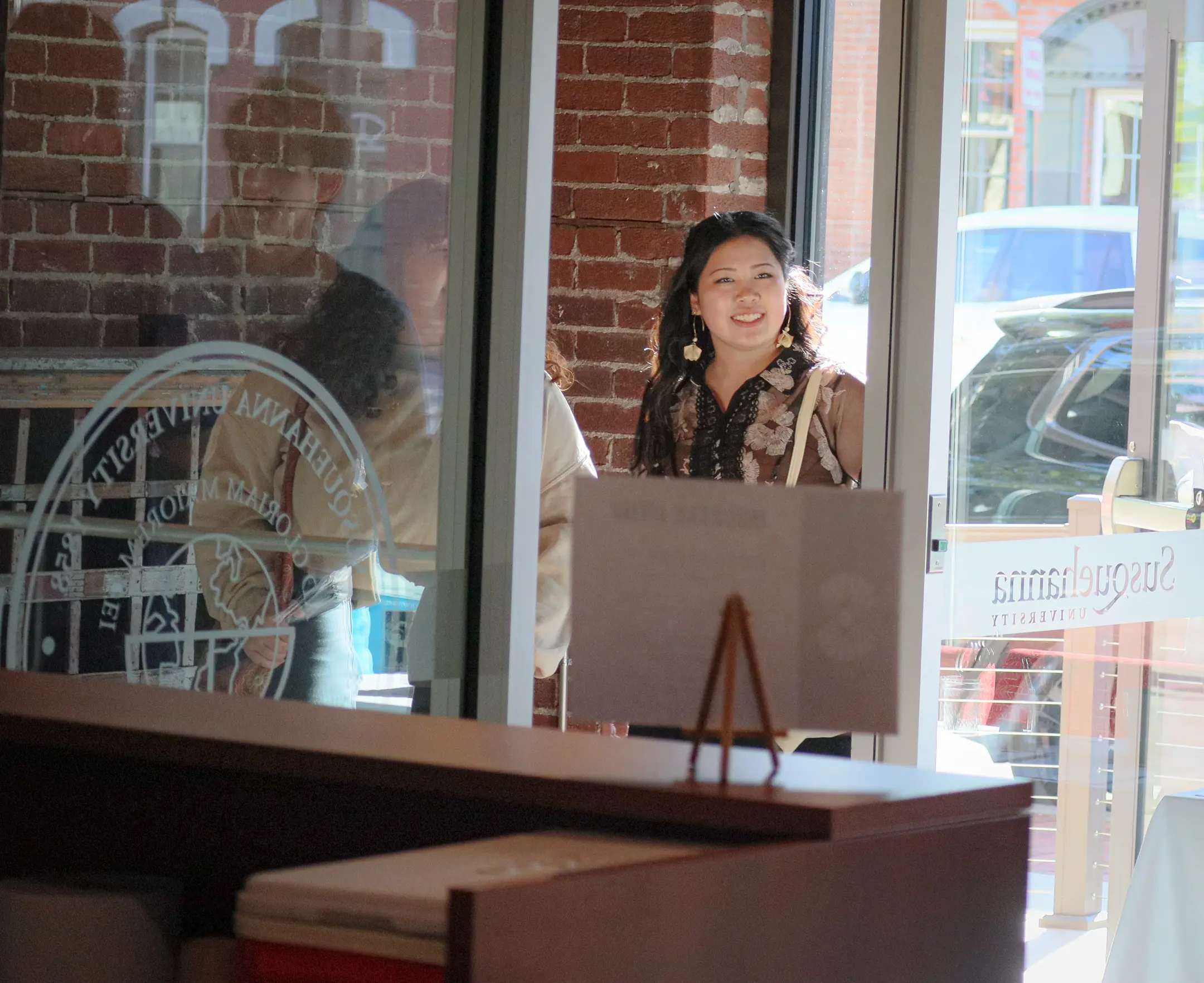 A woman, embodying the essence of campus life, smiles as she walks into a sunlit building with brick walls and glass doors. Her patterned blouse and earrings catch the light. Behind her, another person is partially visible, while in the background a parked car and window signage complete the scene.