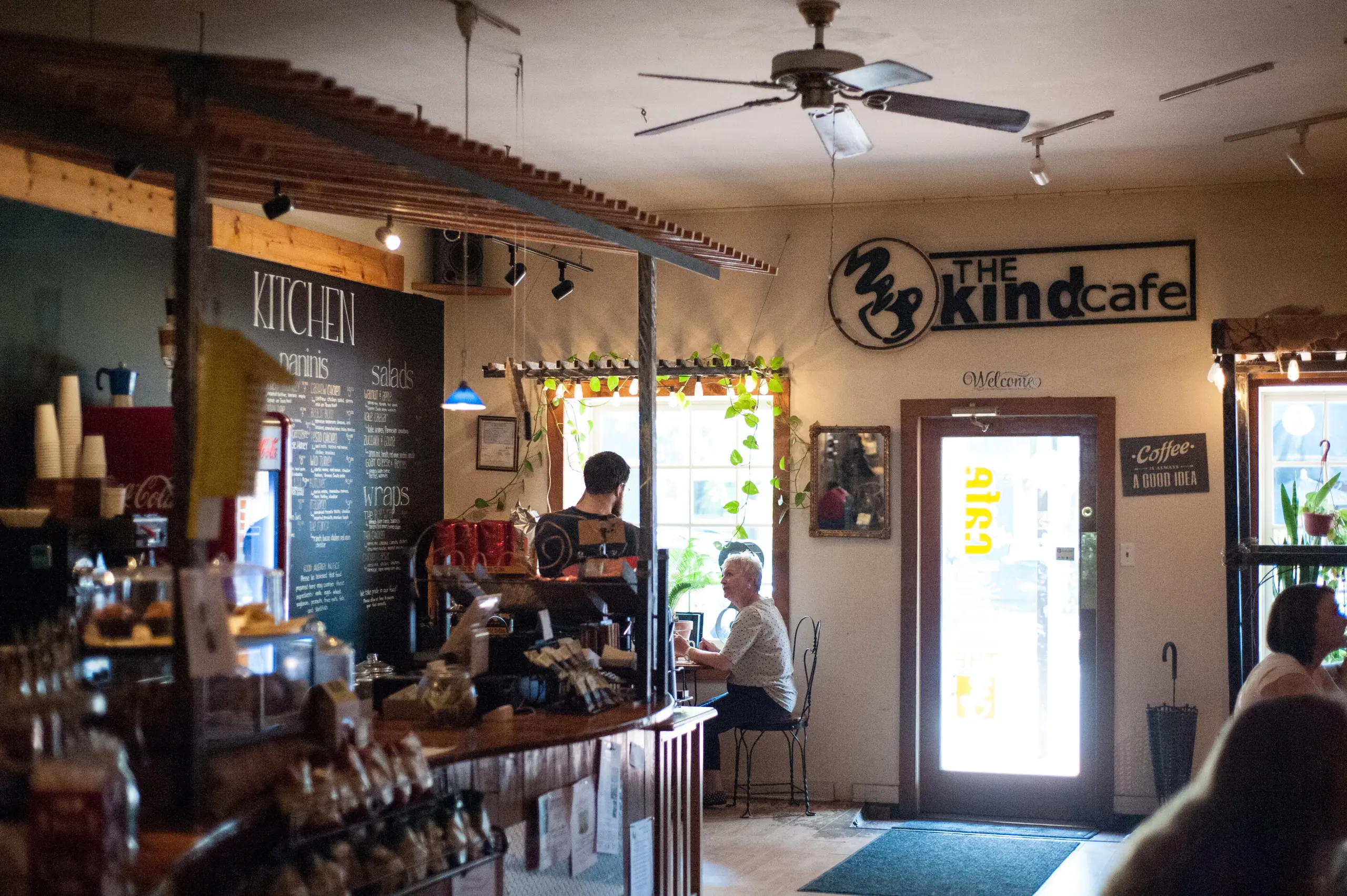 The Kind Cafe offers a cozy retreat with dim lighting and lush plants, perfect for taking a break from campus life. A large menu graces the wall beside the counter, while students mingle at tables, enjoying conversation in this warm and welcoming atmosphere.