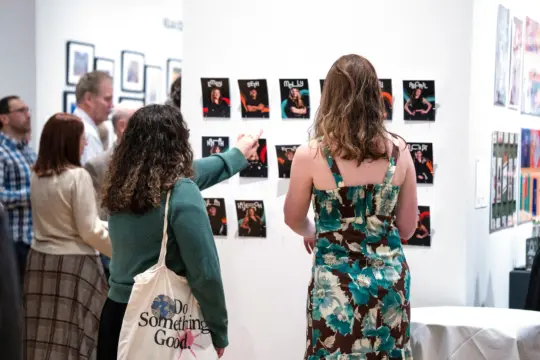 At the Lore Degenstein Gallery, a group observes photographs on display. In the foreground, one person points at a photo while another, clad in a floral dress, stands close. The gallery buzzes with activity as several artworks adorn the walls.