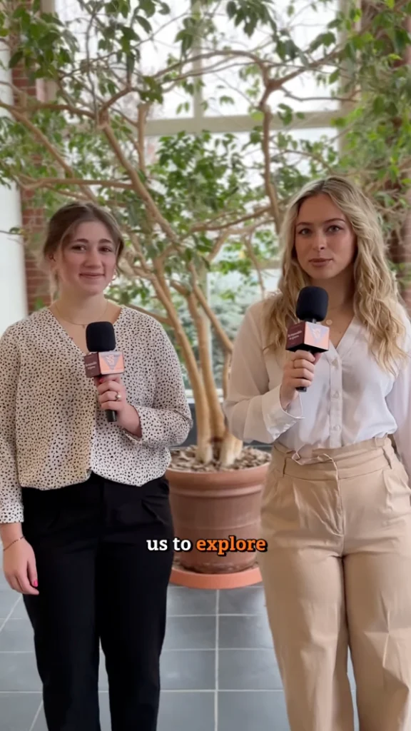 Two people holding microphones stand indoors in front of potted plants. They are wearing business casual attire and appear to be speaking or conducting an interview. Text on the image reads "us to explore.