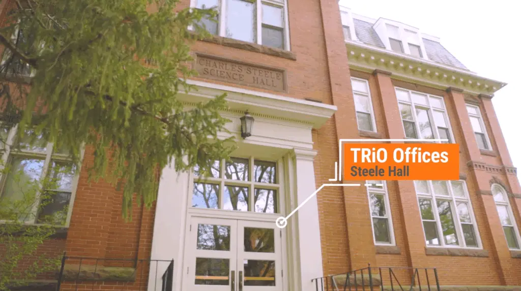 The image shows the entrance to a red brick building labeled "Charles Steele Science Hall." A white arrow directs toward the door with orange text reading "TRiO Offices, Steele Hall." A tree branch is partially visible in the foreground, adding a natural touch.