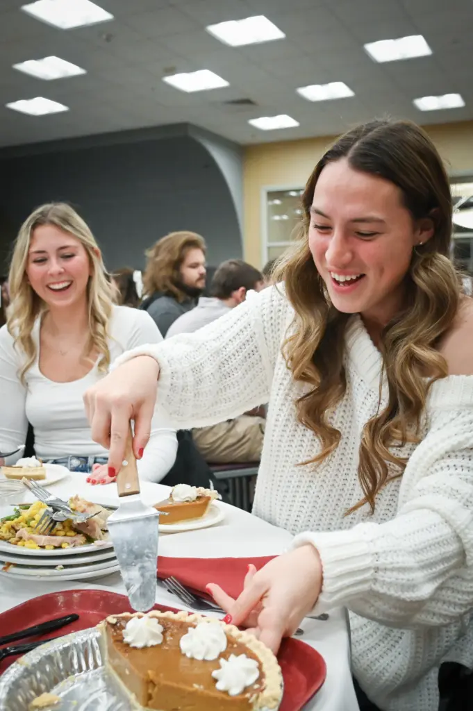 Two young women at a table, embodying the essence of campus life, with one serving a piece of pie. They both smile in matching white tops. The table is adorned with plates of food and a pie, set in a lively dining area bustling with chatter from others in the background.