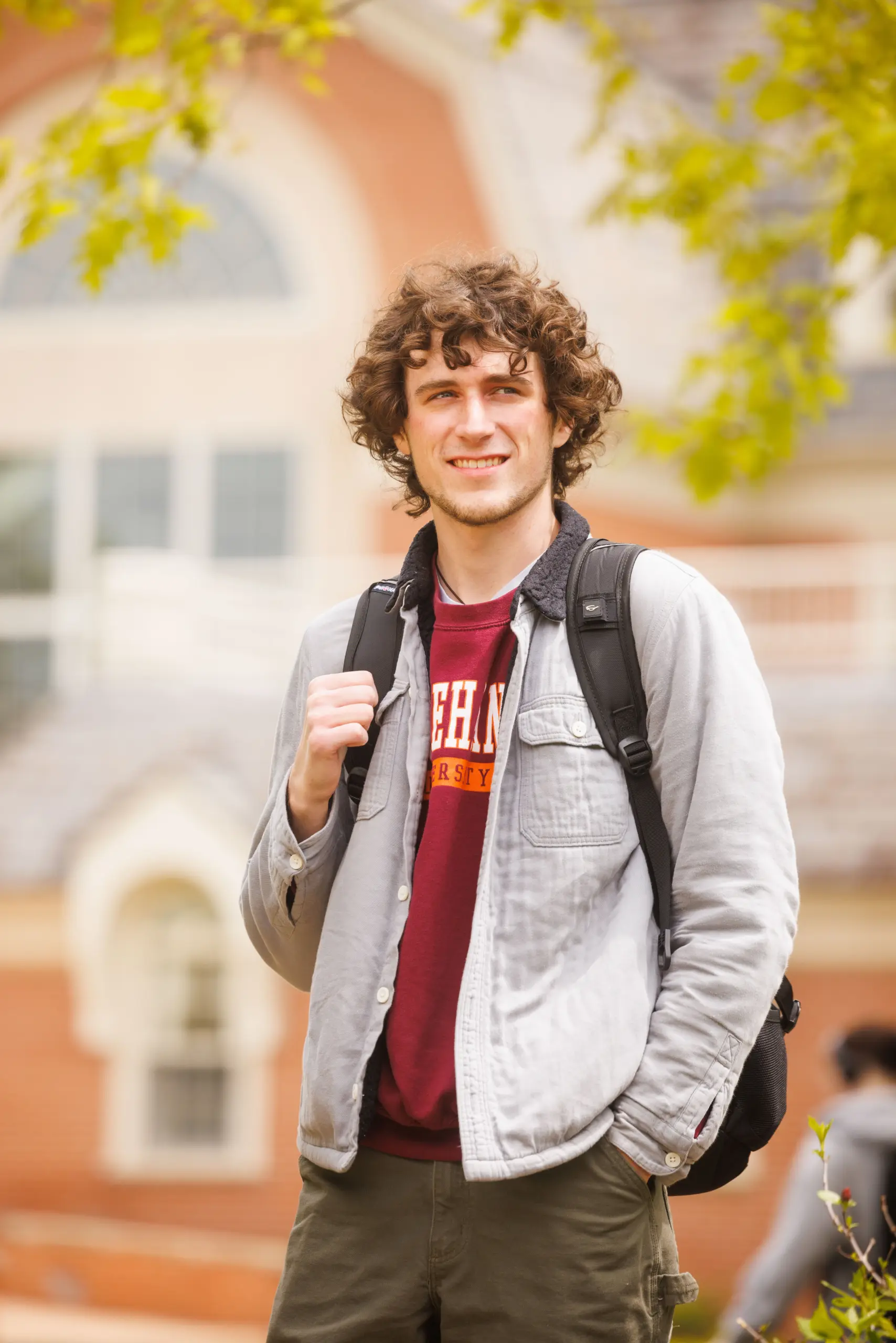 A person with curly hair and a backpack, wearing a gray jacket over a maroon shirt, stands outside in front of a brick building with arched windows. Green leaves are visible in the foreground. They are smiling and looking away from the camera.